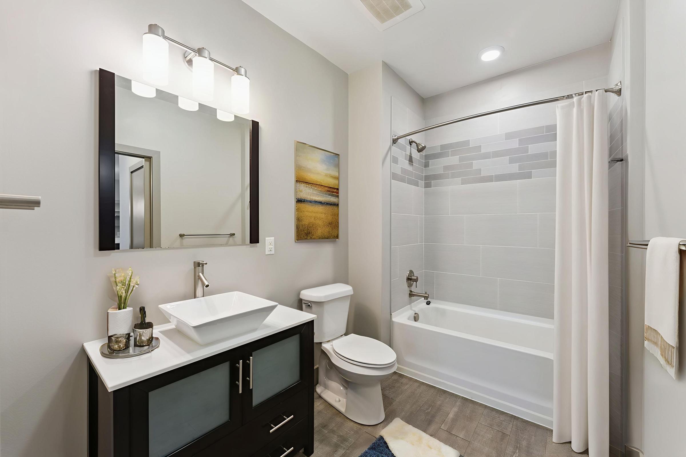 A modern bathroom featuring a white bathtub with a shower curtain, a sleek vanity with a vessel sink, and a wall-mounted mirror. Neutral-colored walls and flooring create a clean, contemporary look. A piece of artwork and decorative items add a touch of style to the space.