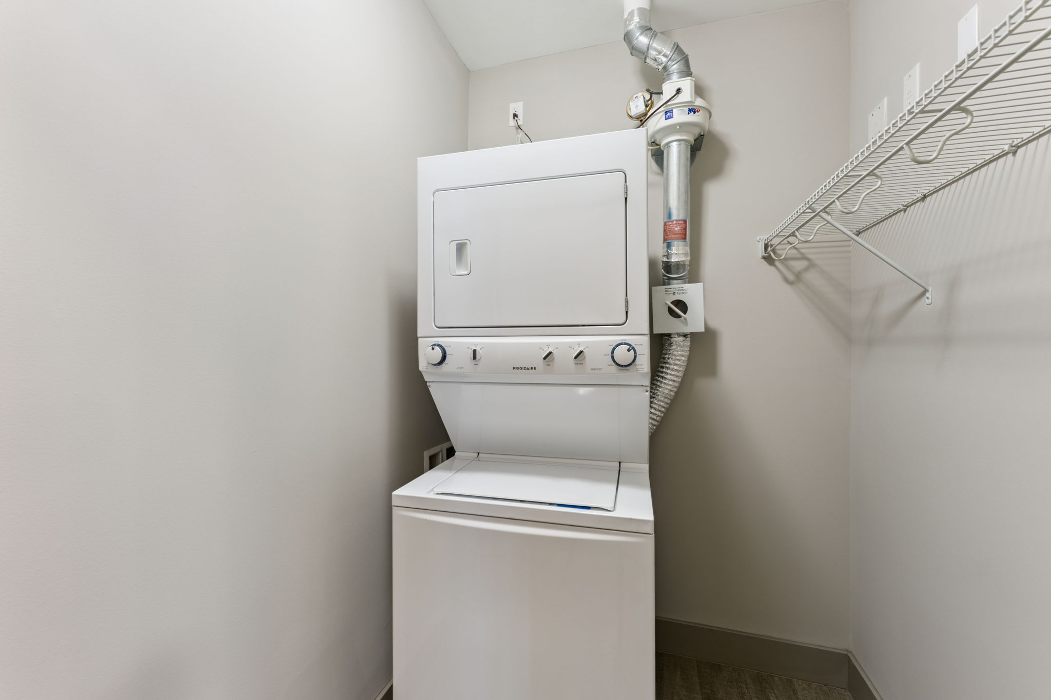 A stacked washer and dryer unit in a small laundry room. The appliances are white, with a metal vent pipe attached. The walls are painted light gray, and there is a wire shelf above the unit. The floor features a neutral-colored flooring material.