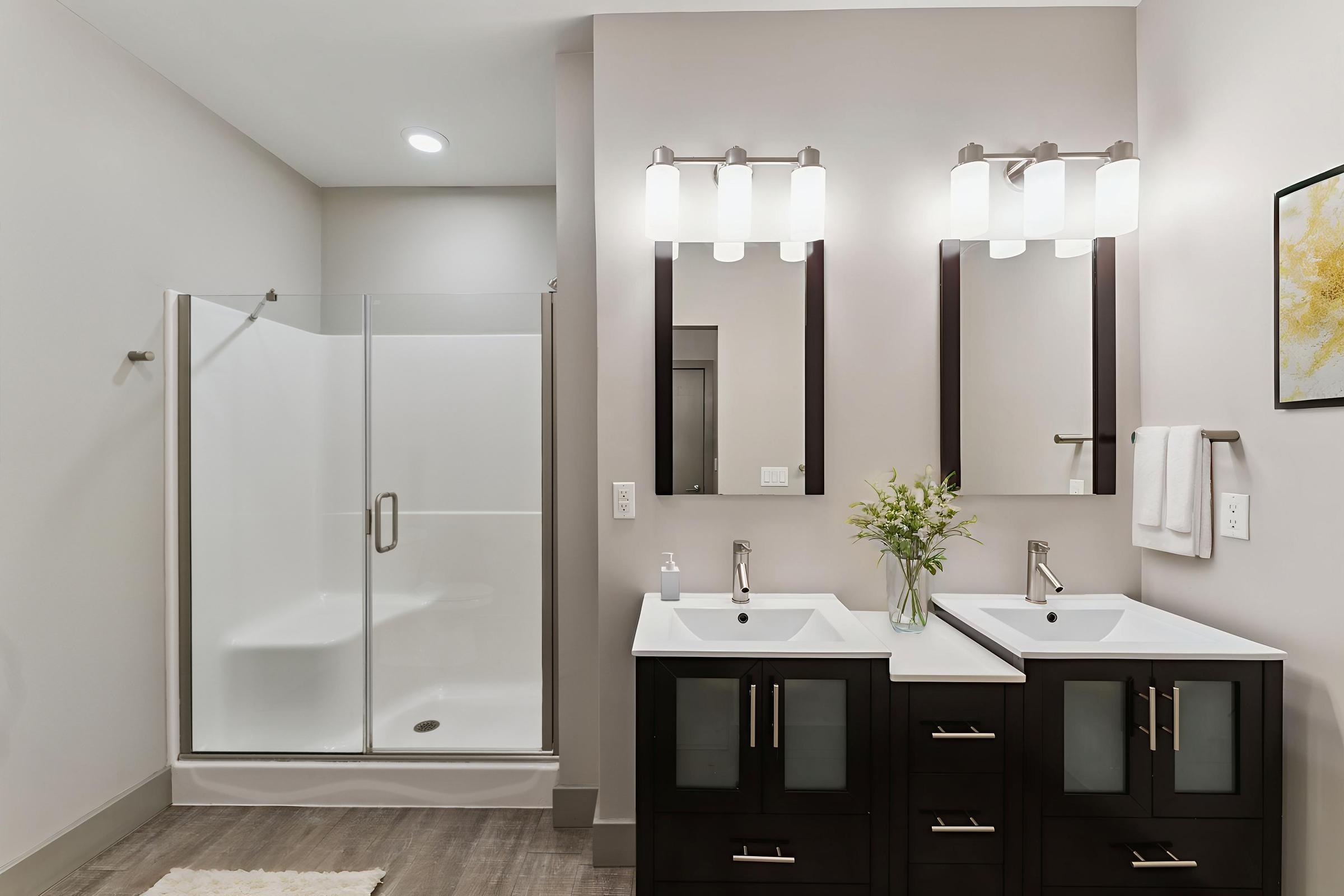 Modern bathroom with a double vanity featuring two sinks and mirrors above. There is a glass shower enclosure on the left and neutral-colored walls. The decor is minimalistic, with a small vase of flowers on the counter and a piece of art on the wall for a touch of color. Soft lighting enhances the contemporary design.
