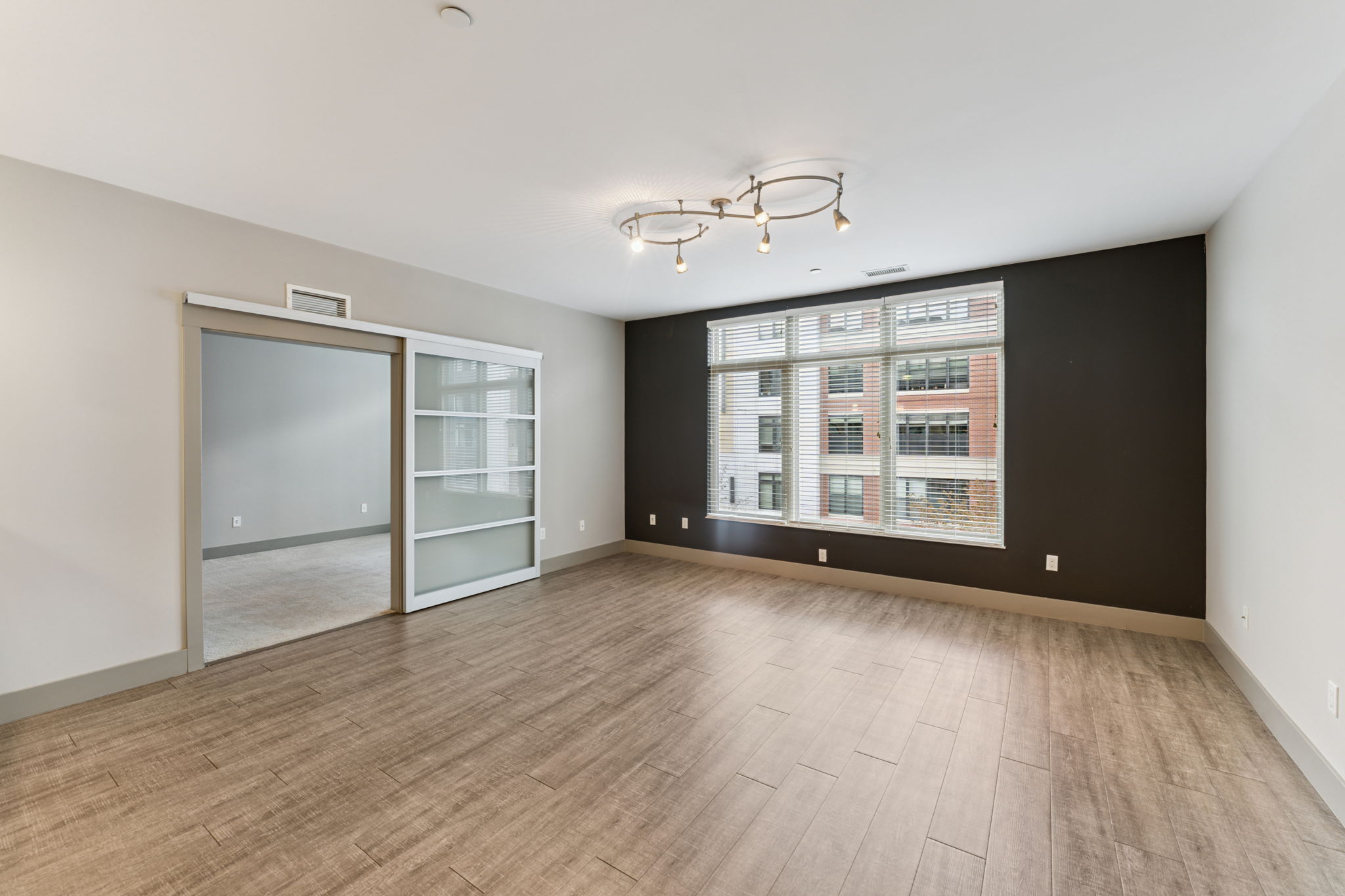 A spacious, modern living room featuring a large window with natural light, a dark accent wall, and laminate flooring. There is a glass-door closet on the left and track lighting on the ceiling, enhancing the contemporary design of the space.