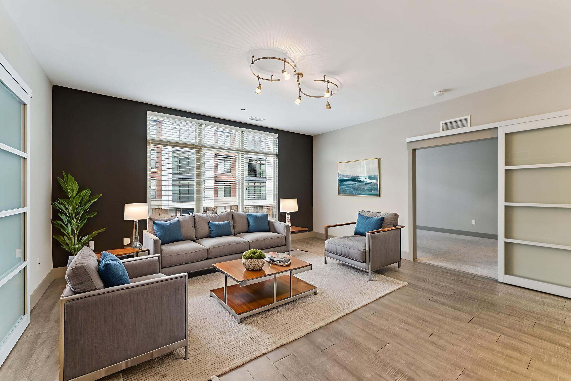 Modern living room featuring a comfortable gray sofa with blue cushions, two armchairs, and a coffee table on a light rug. Natural light fills the space from large windows with white blinds. There’s a decorative plant in the corner and a piece of wall art above the couch, with an adjacent open room visible through glass doors.