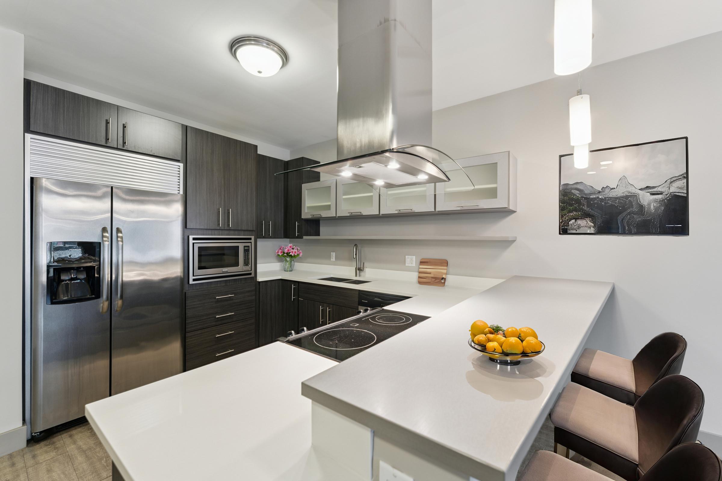 Modern kitchen featuring sleek dark cabinetry, stainless steel appliances including a refrigerator and microwave. The countertop has a built-in stove and a bowl of oranges. One side includes a small dining area with two chairs. A framed picture of a landscape decorates the wall. Natural light fills the space, creating a bright atmosphere.