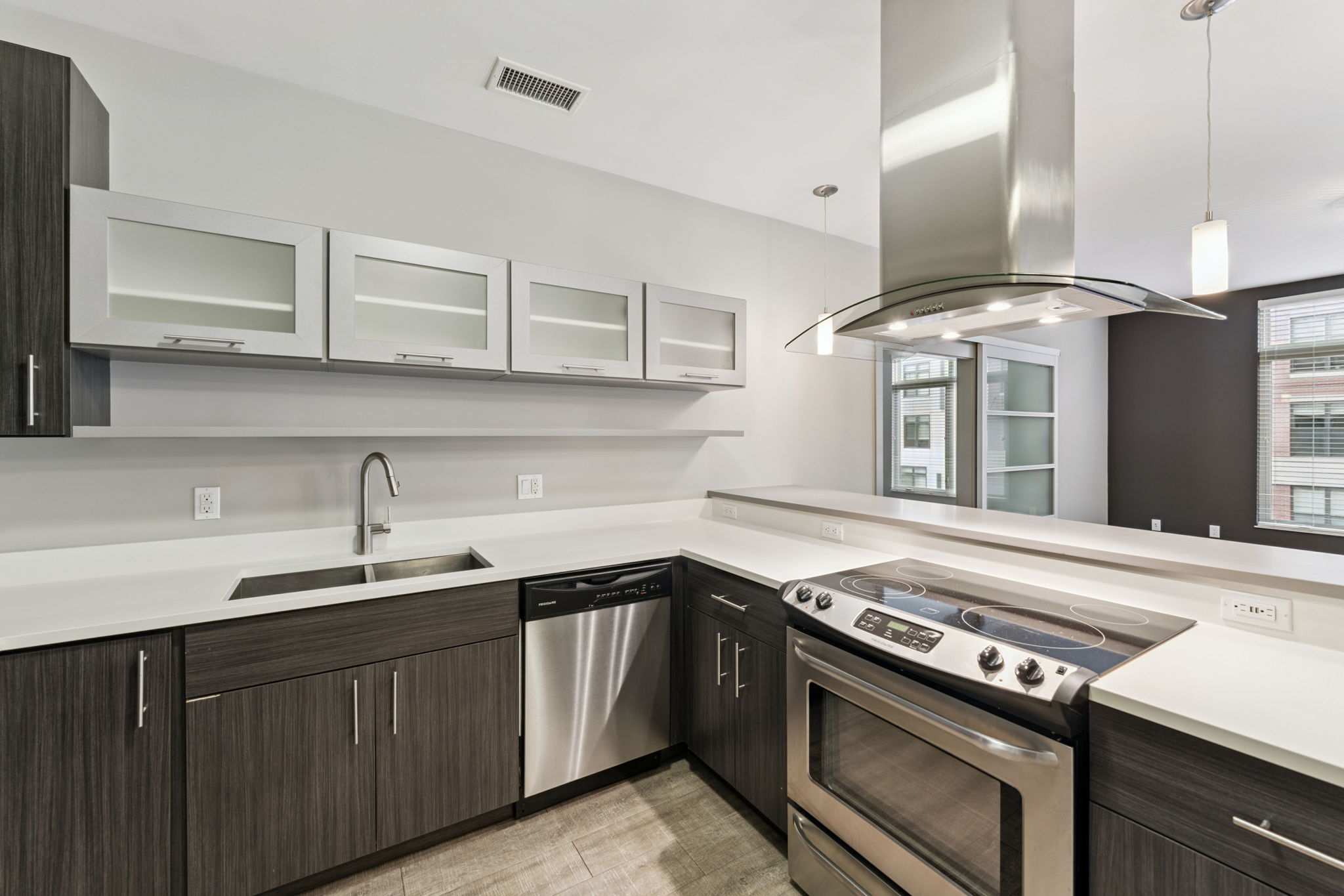 Modern kitchen with dark wood cabinetry and stainless steel appliances. The design features a large stainless steel range hood above a stove, a built-in dishwasher, and an undermount sink. Light-colored countertops complement the space, which includes a window showing an outdoor view.