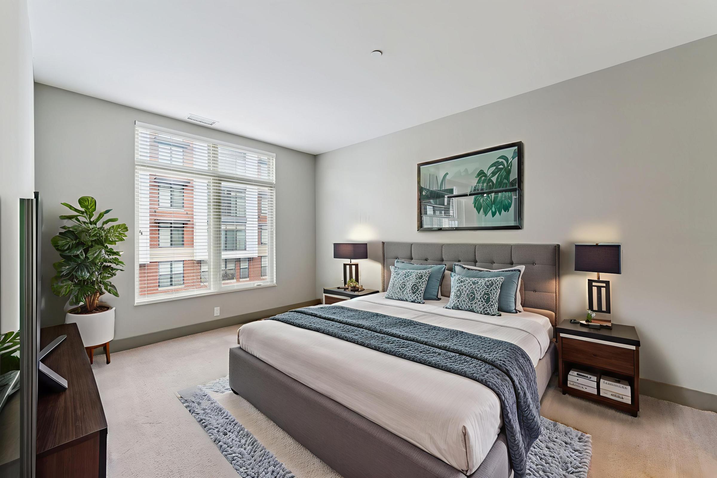 A modern bedroom featuring a large bed with a gray upholstered headboard, decorated with blue and gray pillows. There are two nightstands with lamps on either side, a plant in the corner, and a window with blinds allowing natural light. A piece of wall art is visible above the bed.