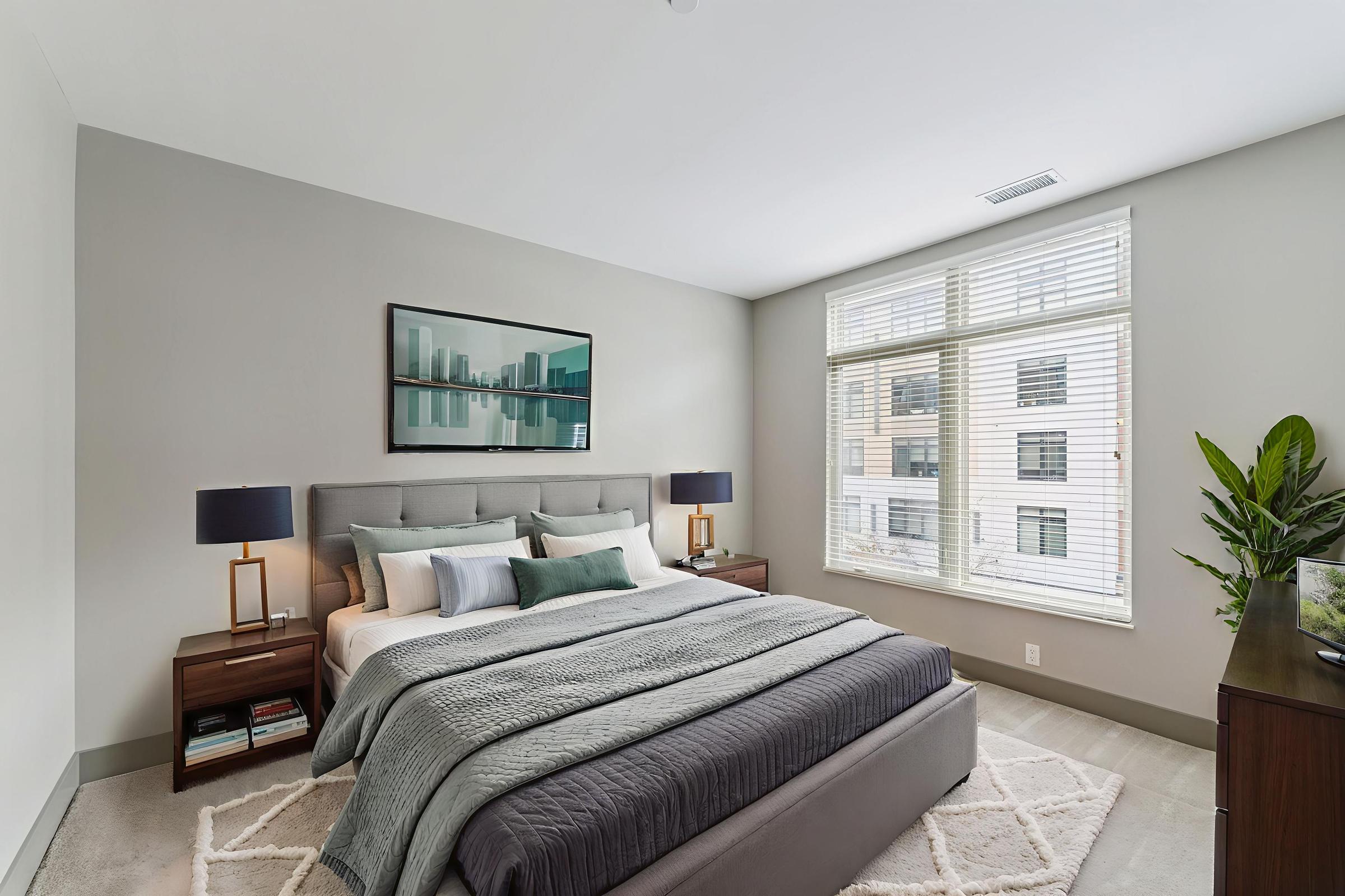 A modern bedroom featuring a large bed with grey bedding and decorative pillows, flanked by stylish bedside lamps. A window with blinds allows natural light, while a small plant adds a touch of greenery. Wall art depicting a cityscape hangs above the bed, and a dresser is present against one wall.