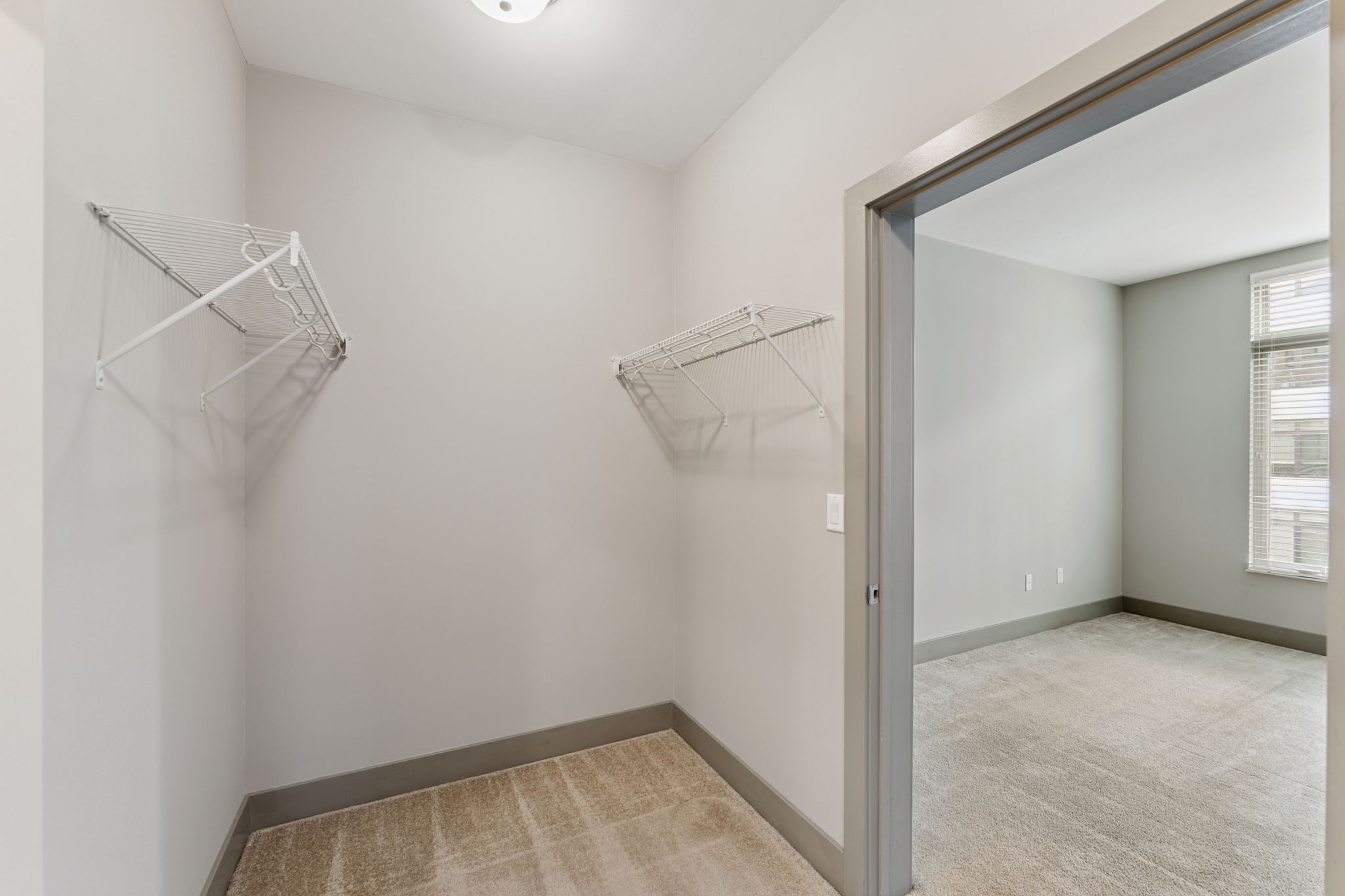 A small, empty room with light gray walls and beige carpet. Two metal wire shelving units are mounted on the wall, and a doorway leads to another room with natural light coming from a nearby window. The overall space appears clean and modern, suitable for storage or as a small closet.