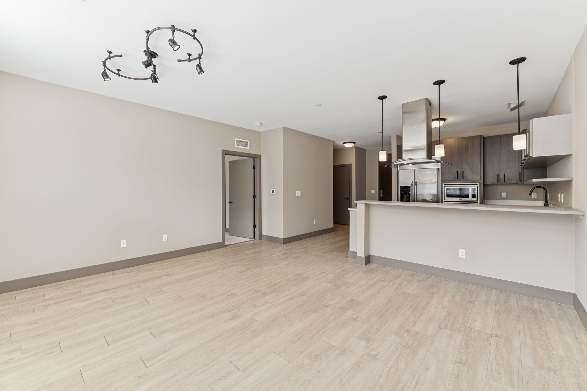 Spacious modern living area with light-colored walls and tiled flooring. The kitchen features a breakfast bar with pendant lighting, stainless steel appliances, and dark wood cabinetry. Open layout with ample natural light, and a doorway leading to another room.