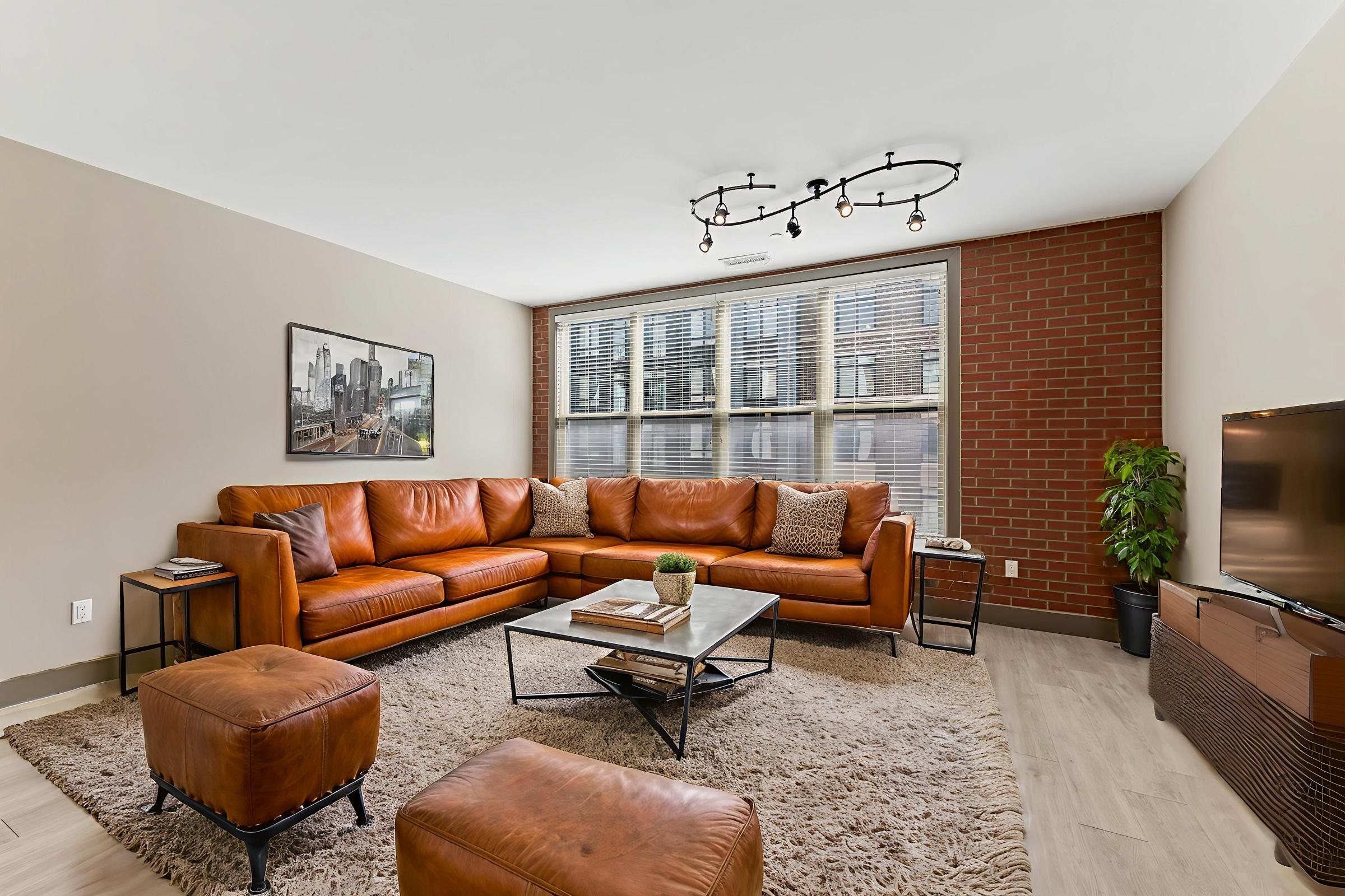 A bright living room featuring a large brown leather sectional sofa, a glass coffee table, and a cozy area rug. Large windows provide ample natural light, while exposed brick and modern decor add warmth. A potted plant and a flat-screen TV complete the inviting space.