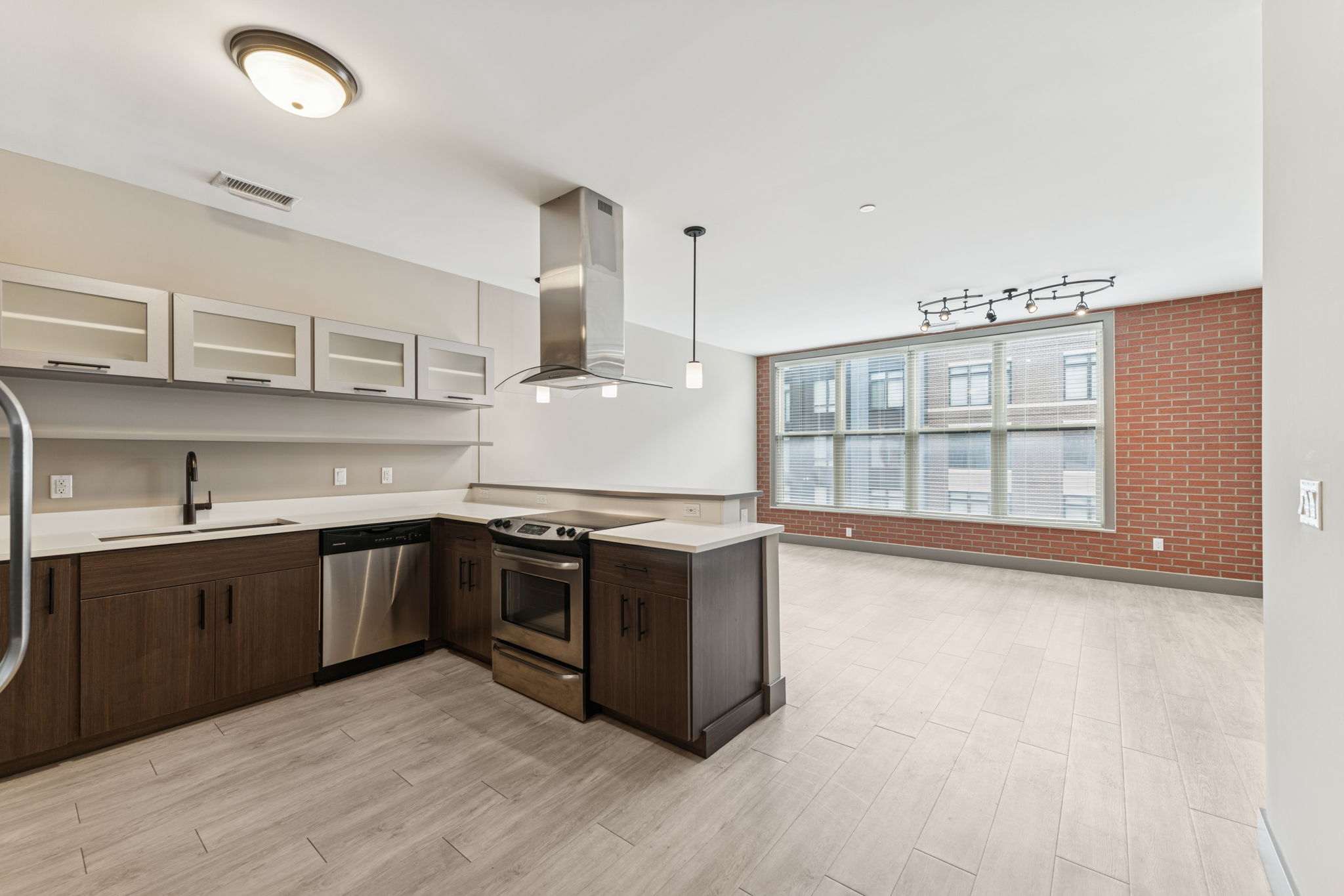Modern open-concept kitchen and living area featuring sleek dark cabinetry, stainless steel appliances, and light-colored tile flooring. Large windows provide natural light, and exposed brick accents add character to the space. A range hood is installed above the oven, enhancing the contemporary design.