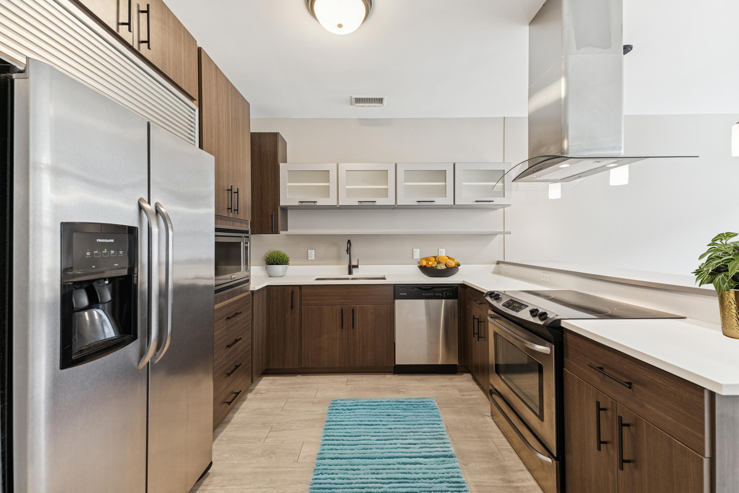 Modern kitchen featuring stainless steel appliances, including a refrigerator and oven. Light wood cabinetry and a white countertop provide a sleek look. There's a blue rug on the floor, and a plant decorates the countertop. Open shelving displays items, and the space is well-lit with overhead fixtures.