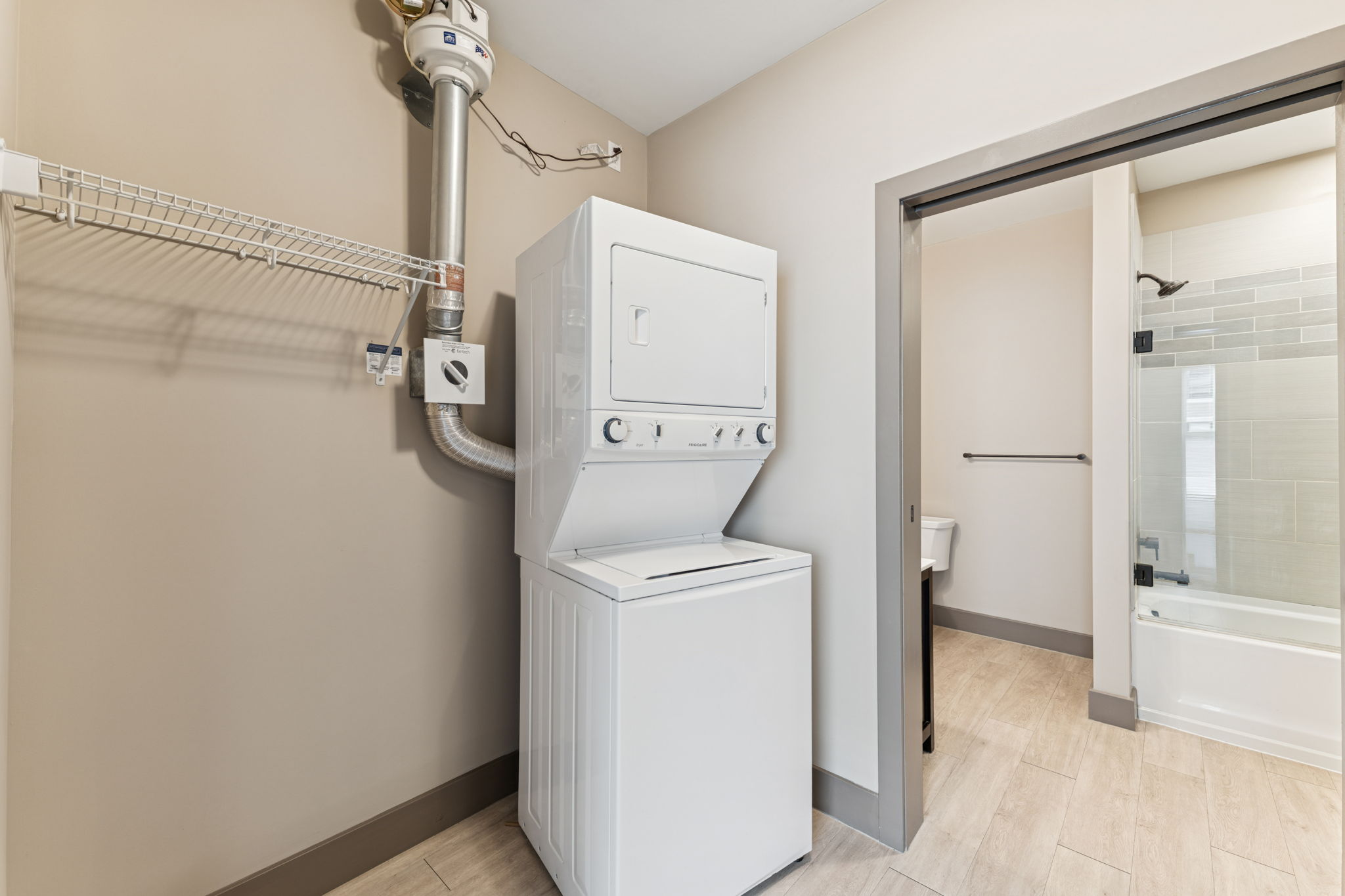 A compact laundry area featuring a stacked washer and dryer unit. The space is modern with light-colored walls and wood-look tile flooring. A metal shelf is mounted on the wall for storage, and a bathroom area is visible through an open door on the right, showcasing a shower and toilet.