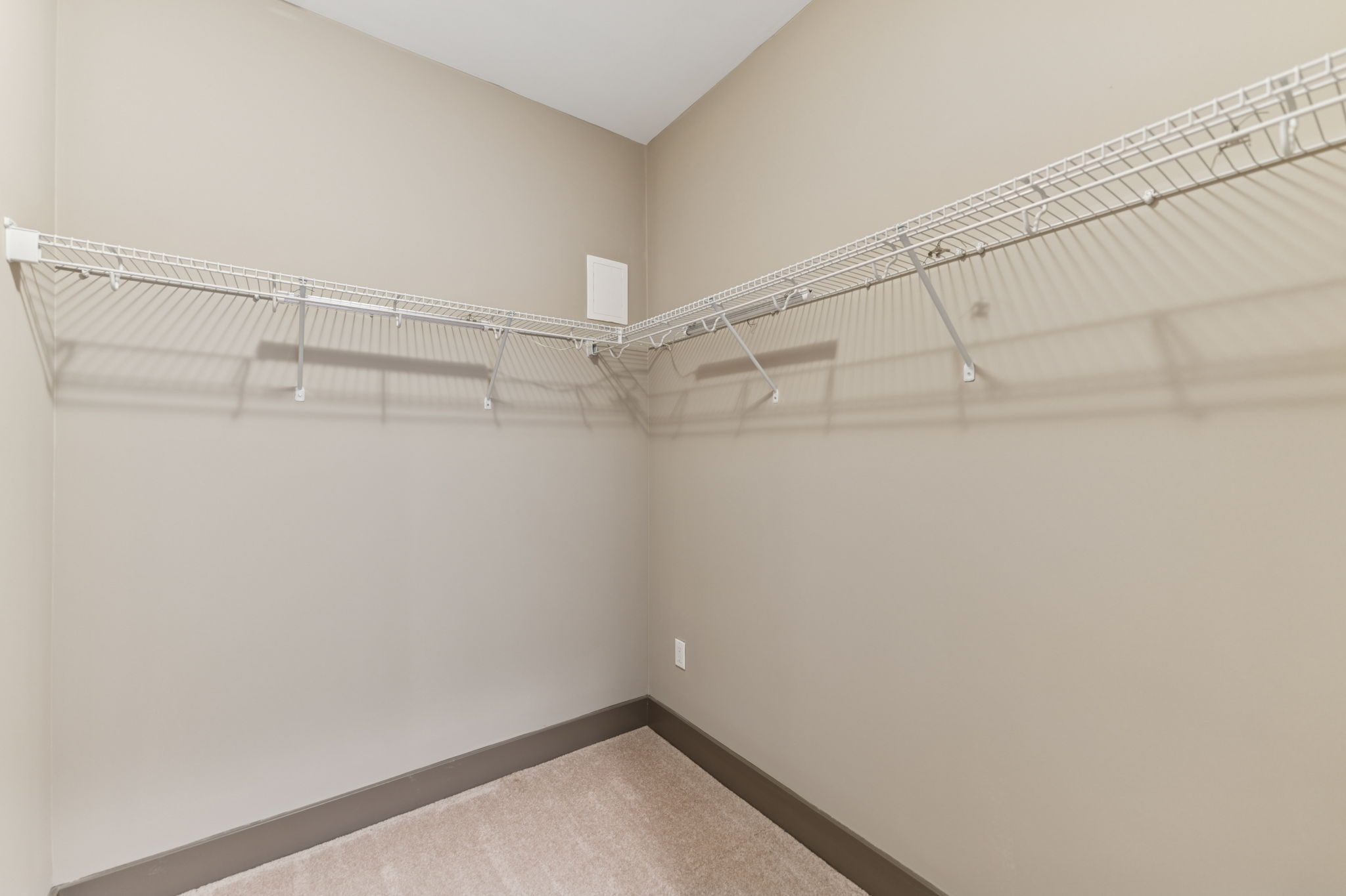 A spacious, empty closet featuring wire shelving along the walls and a soft carpeted floor. The walls are painted a light beige, creating a neutral backdrop, while the shadows of the shelves add depth to the uncluttered space.