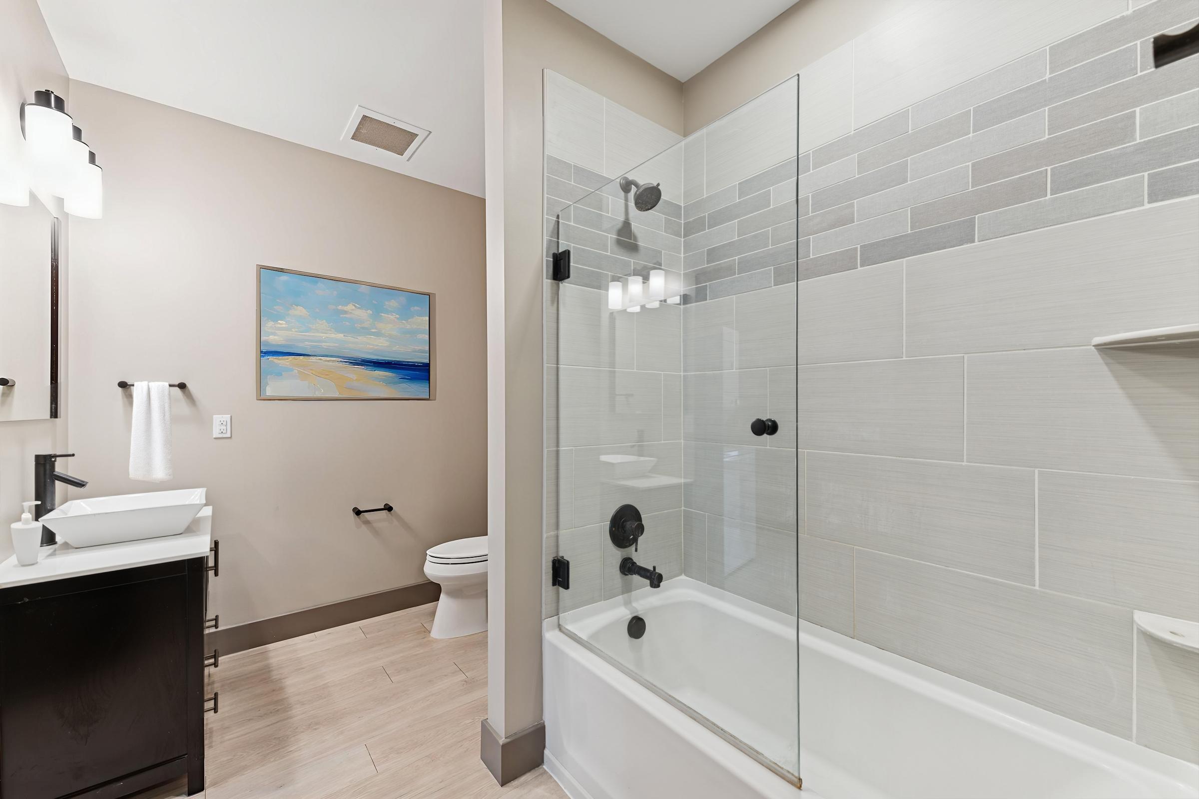 Modern bathroom featuring a glass shower enclosure, a white bathtub, and stylish tile work. Includes a sleek vanity with a vessel sink, a modern toilet, and a framed beach landscape painting on the wall. The decor emphasizes a clean, contemporary aesthetic with neutral colors.