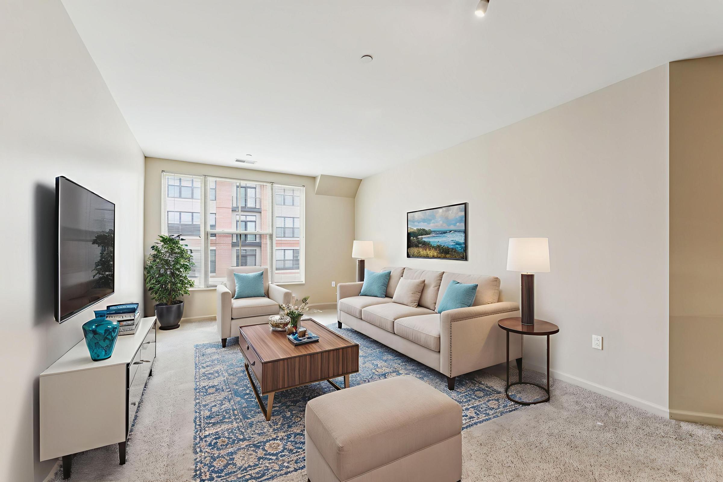 A modern living room featuring a beige sofa with blue accent pillows, a wooden coffee table, and a television mounted on the wall. Two lamps flank the sofa, and a potted plant adds greenery. A large window lets in natural light, and a patterned area rug enhances the cozy atmosphere.