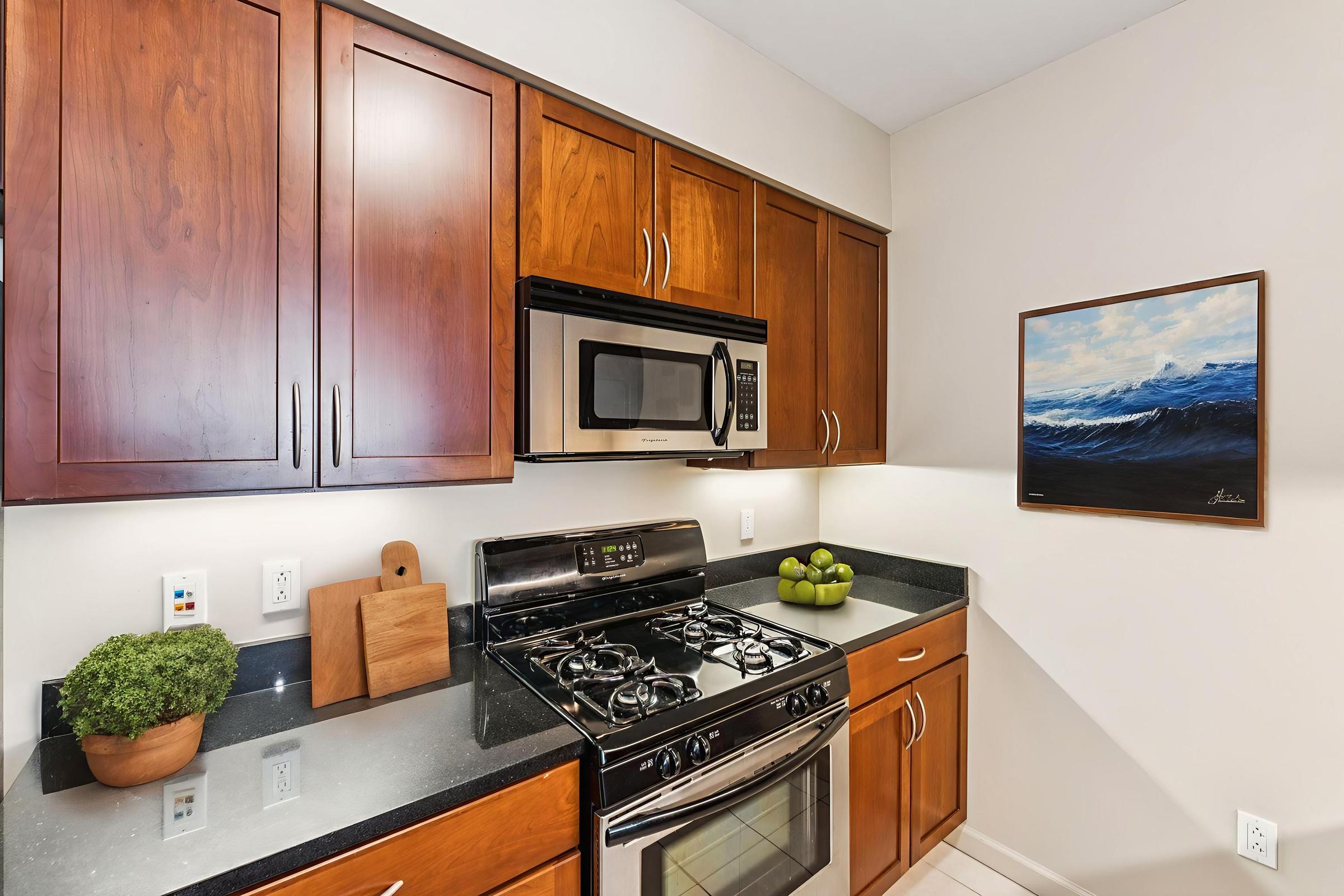 A modern kitchen featuring wooden cabinets, a stainless steel microwave, and a gas stove. The countertop is dark with a small green plant and a bowl of limes. A painting of ocean waves hangs on the wall, adding a touch of color to the space. The overall design is sleek and contemporary.