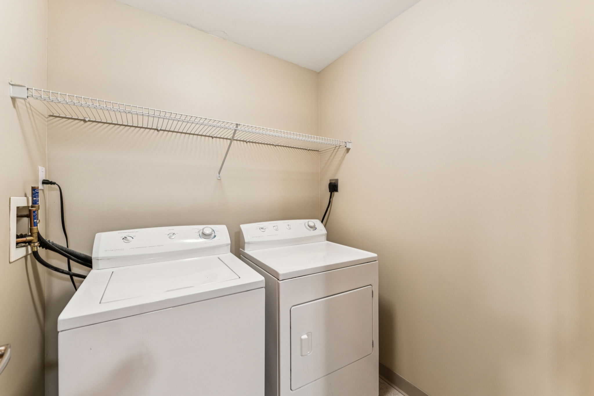 A clean laundry room featuring two white washing machines side by side, placed against a beige wall. There is a wire shelf above the machines, and the flooring is neutral-toned. The space appears well-organized and well-lit.
