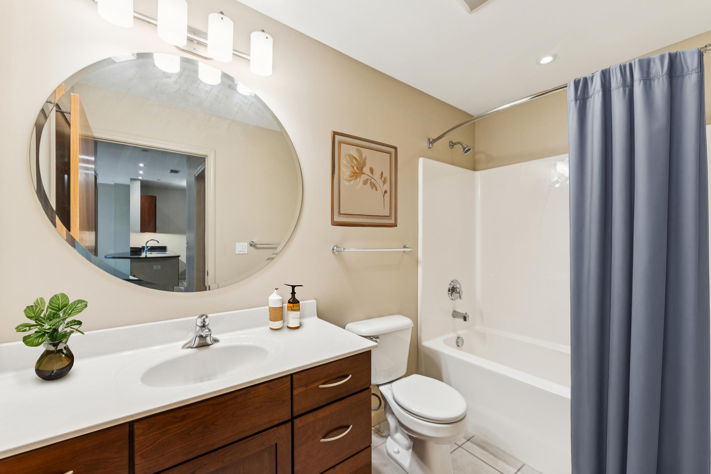 A modern bathroom featuring a round mirror with bright lighting above a white sink. Below the sink, there are wooden cabinets. A white bathtub with a shower curtain is on one side, and a toilet is adjacent. A small green plant and two bottles are on the sink. Neutral wall colors enhance the space.