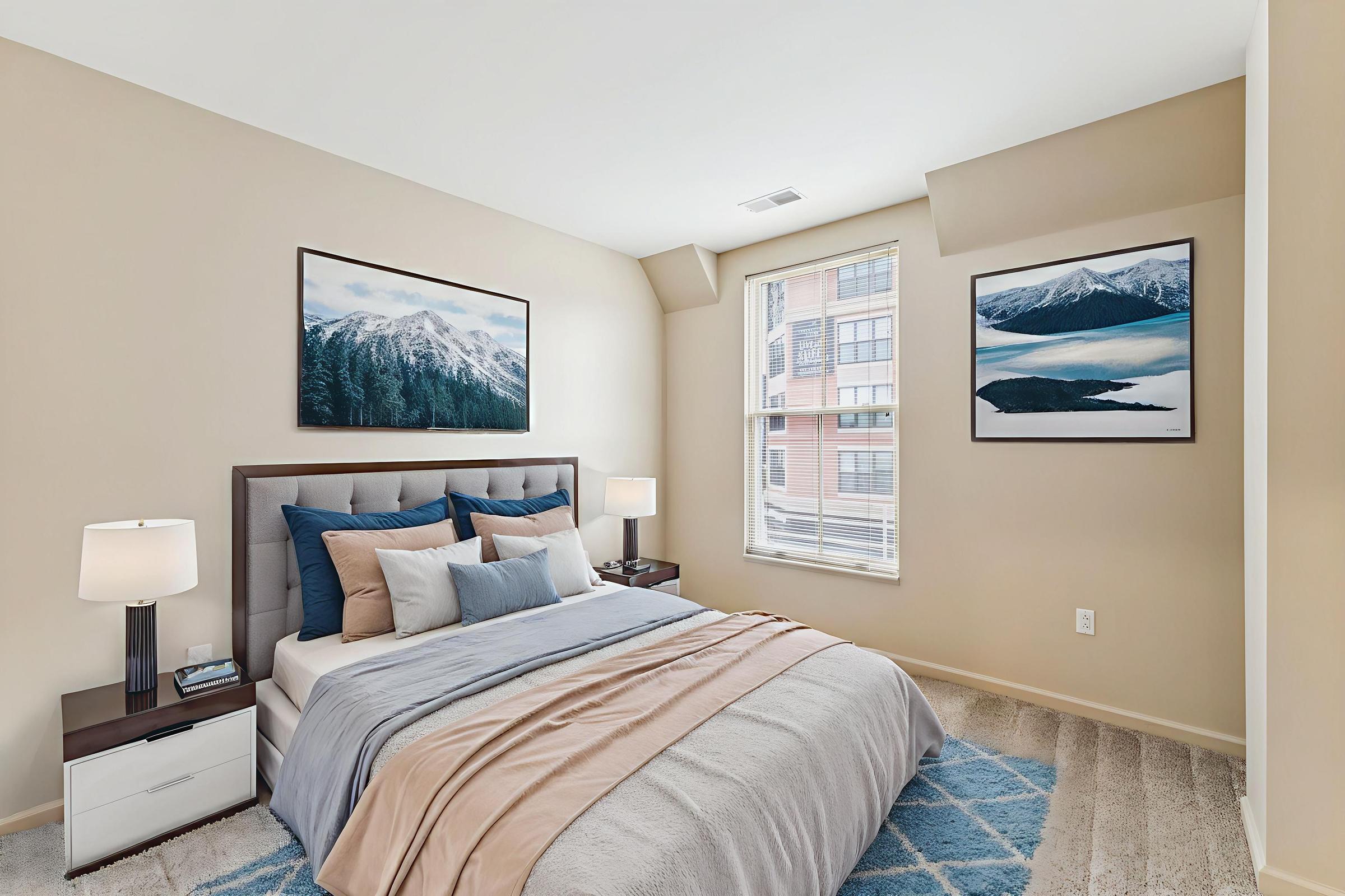 Cozy bedroom featuring a neatly made bed with beige and blue bedding, two bedside lamps, and a modern wooden headboard. The walls are painted in a soft beige, and there are two landscape paintings on the walls. A window allows natural light to brighten the room, showcasing a view of the city.