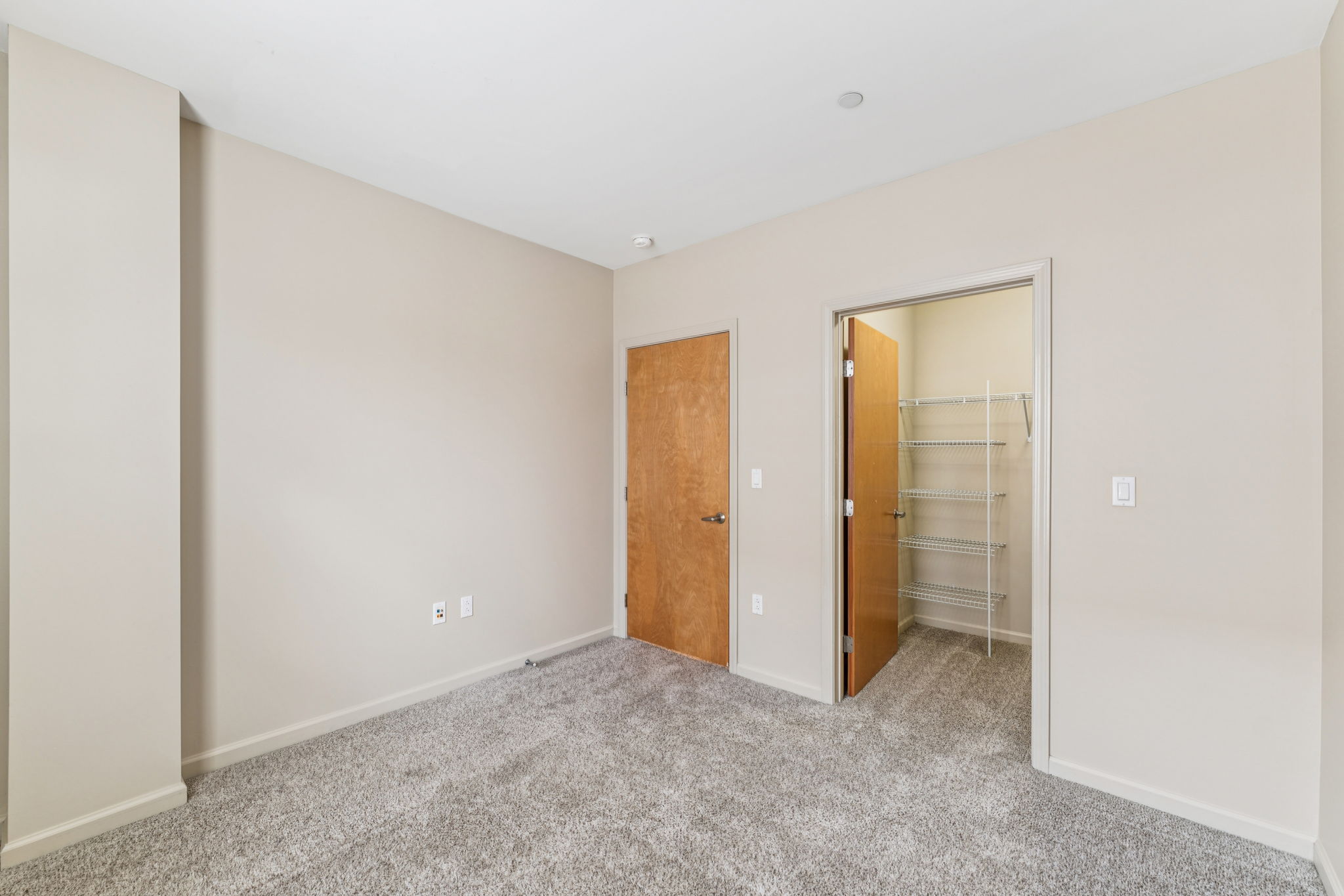 A vacant room featuring light beige walls and a plush, light gray carpet. There is a closed wooden door on one side, with a doorway leading to a closet with wire shelving. The room is well-lit and has a minimalist appearance, making it look spacious and inviting.