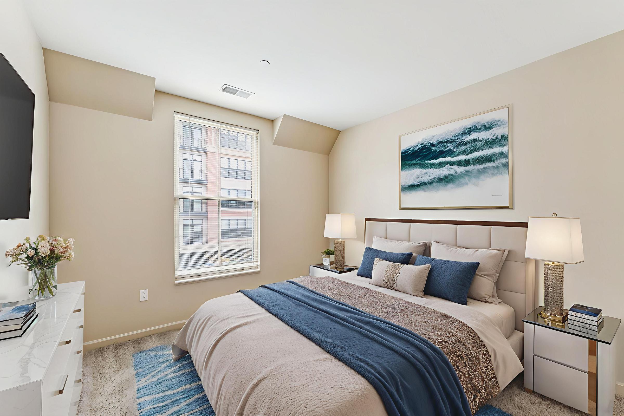 A cozy bedroom featuring a queen-sized bed with a light beige blanket and blue accents. There are two bedside tables with lamps, a modern television mounted on the wall, and a large window showing an urban view. A framed ocean-themed artwork complements the decor.