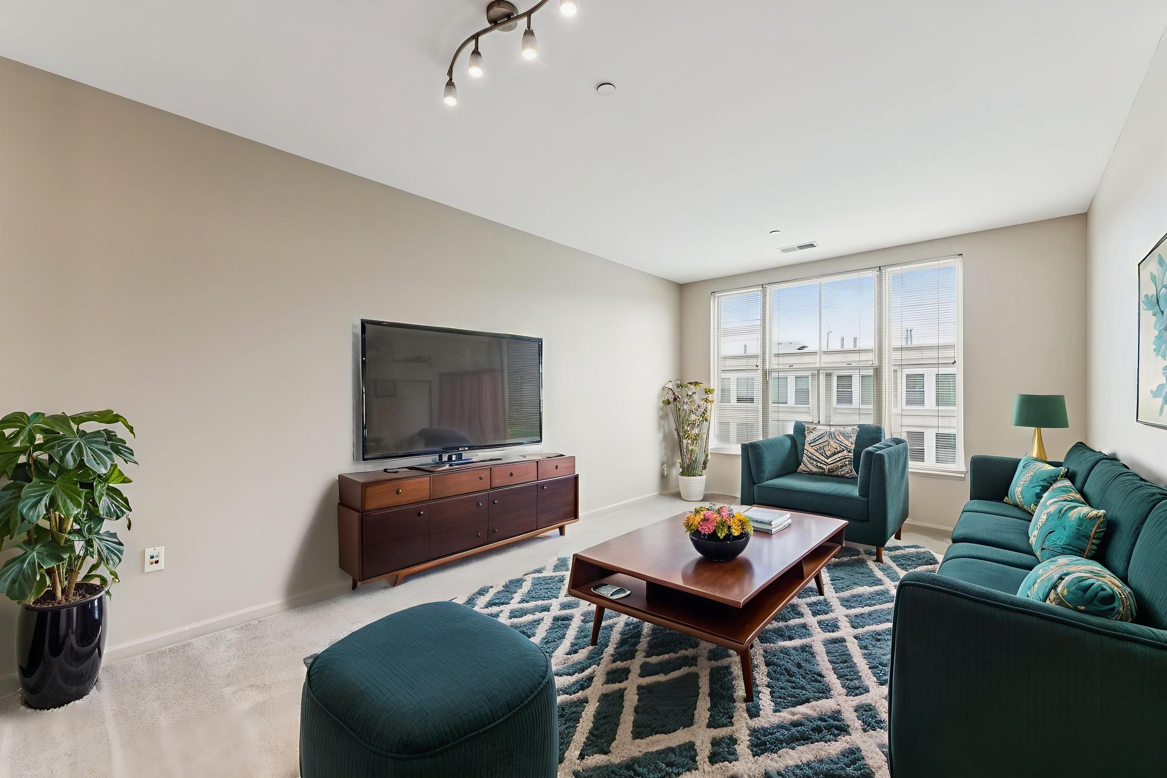 A modern living room featuring a teal sofa, a wooden coffee table, and a large flat-screen TV against a light beige wall. There’s a potted plant in the corner and decorative floral artwork on the wall. Natural light streams in through large windows, illuminating the space with a cozy atmosphere.