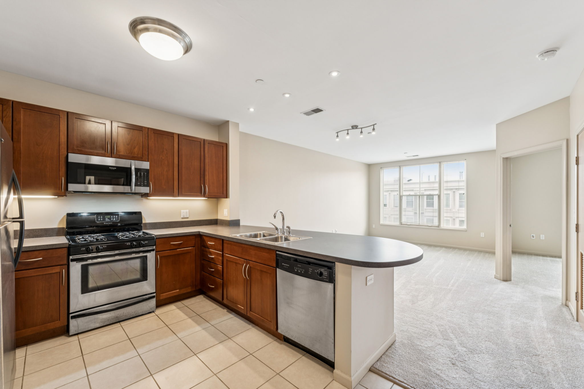 Modern kitchen with wooden cabinets, stainless steel appliances including a gas stove and microwave, and a dishwasher. The kitchen is open to a bright living area with light-colored carpet and a large window providing natural light. The layout suggests a spacious and contemporary living space.