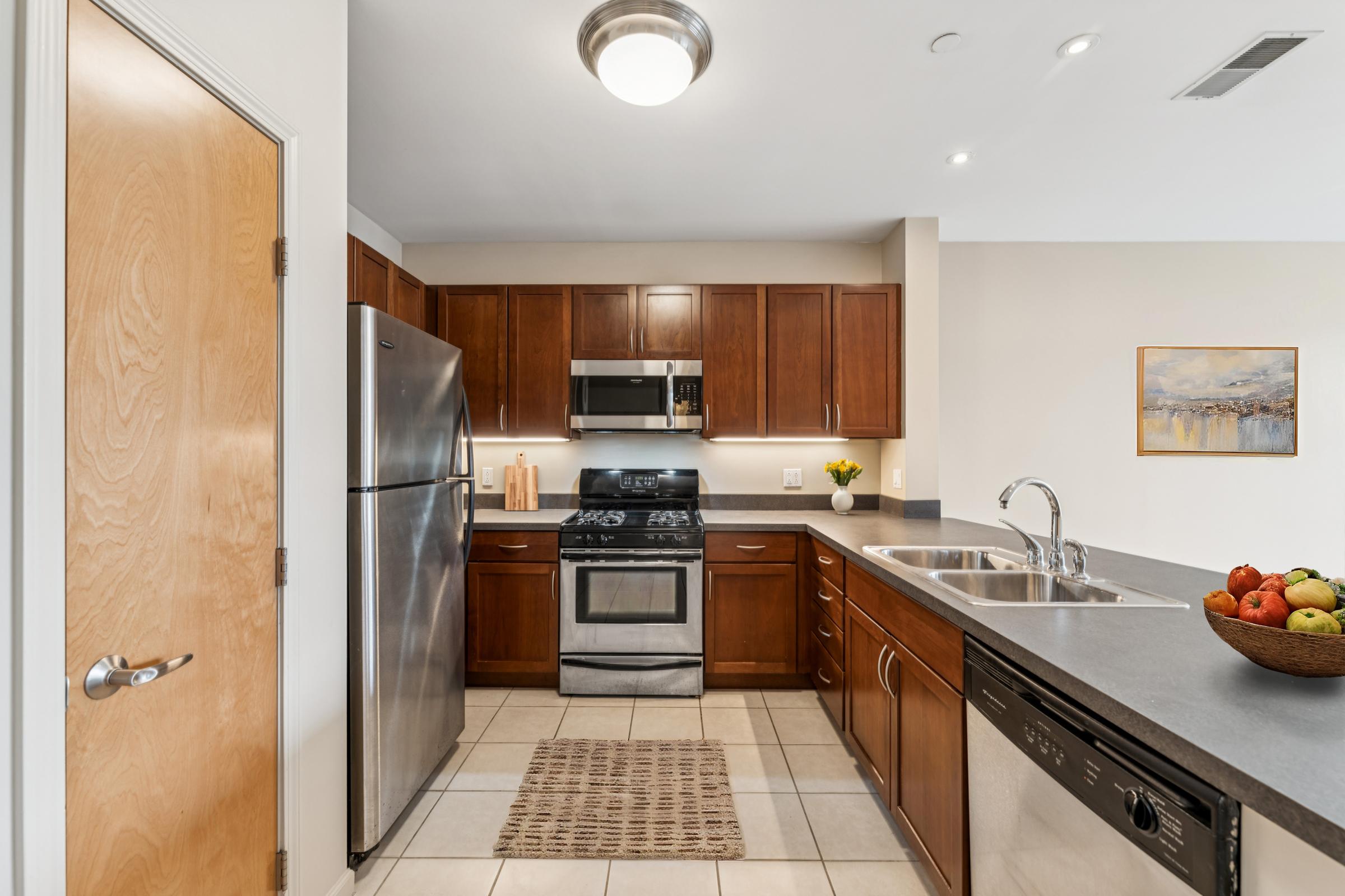 Modern kitchen featuring stainless steel appliances, including a refrigerator, stove, and microwave. Dark wooden cabinets line the walls, and there’s a countertop with a sink. A woven rug is placed on the tiled floor, and a bowl of colorful fruit sits on the countertop, adding a touch of warmth. A small painting is hung on the wall.