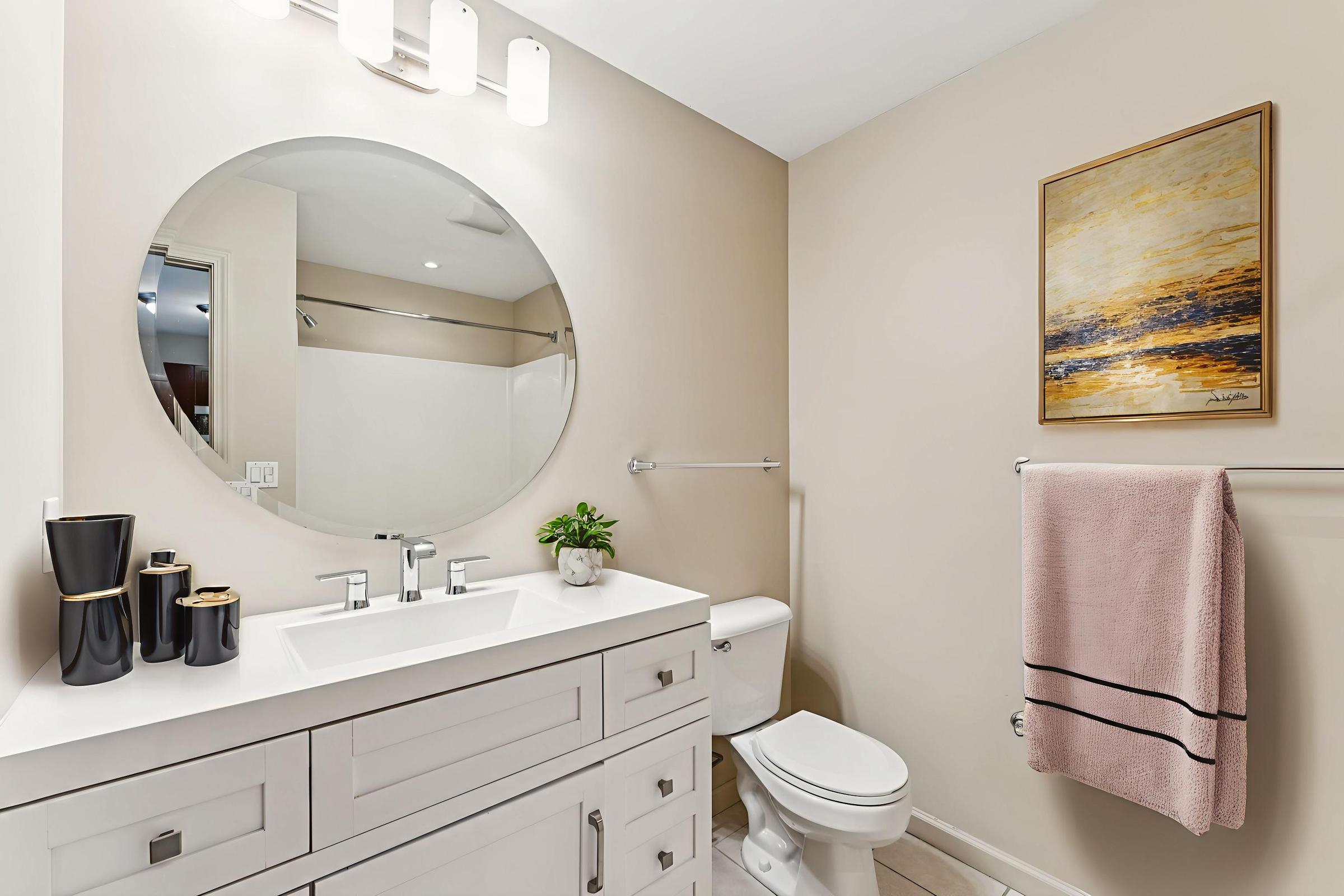A modern bathroom featuring a circular mirror above a sleek white sink with a minimalist vanity. To the left, there is a black and white decorative vase and a small green plant. A light pink towel hangs on a rack, and the walls are painted a soft beige. A piece of abstract art in warm colors hangs beside the mirror.