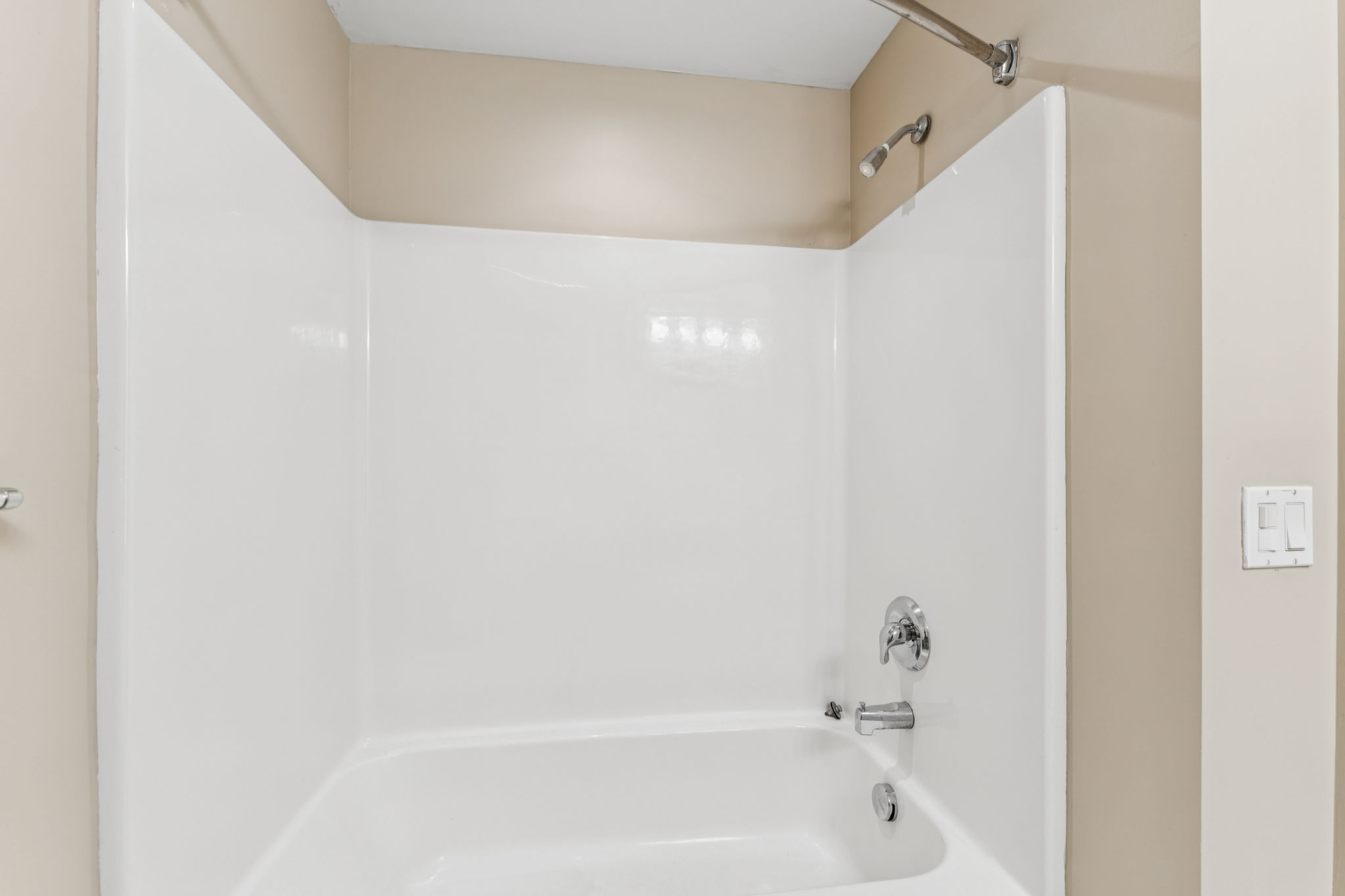 A clean, modern shower stall featuring a white bathtub and walls. The showerhead and faucet are visible, with a simple design. The surrounding walls are painted a light beige, contributing to a bright and open feel in the space. A single light switch is located on the right side wall.