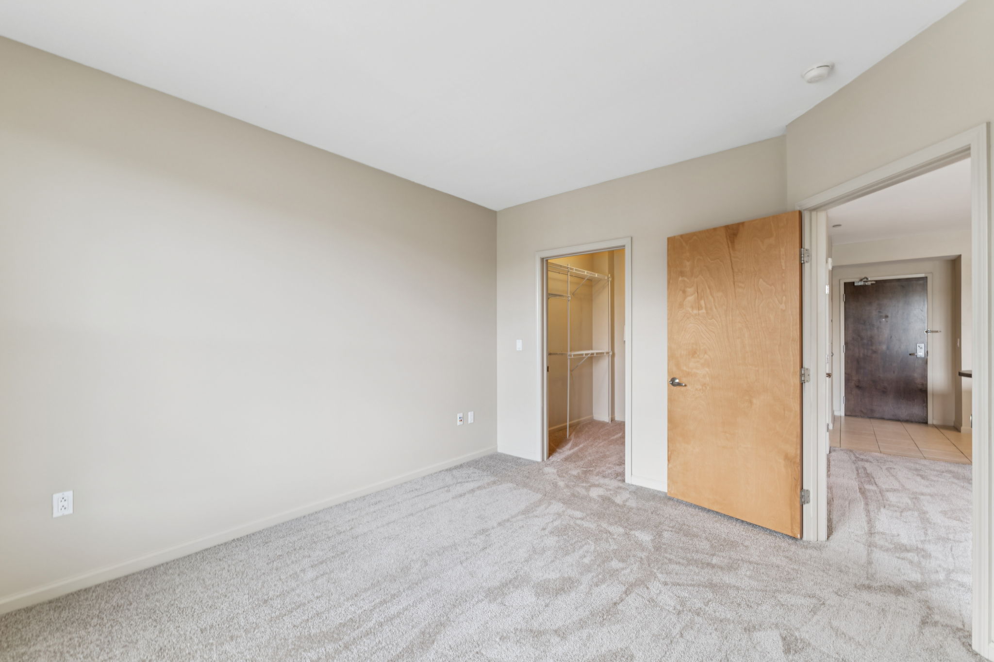 Empty bedroom with light beige walls and carpet. An open door leads to a closet space on the left, and another door on the right opens to a hallway. There’s a window visible on the far wall, and the overall atmosphere is bright and spacious.
