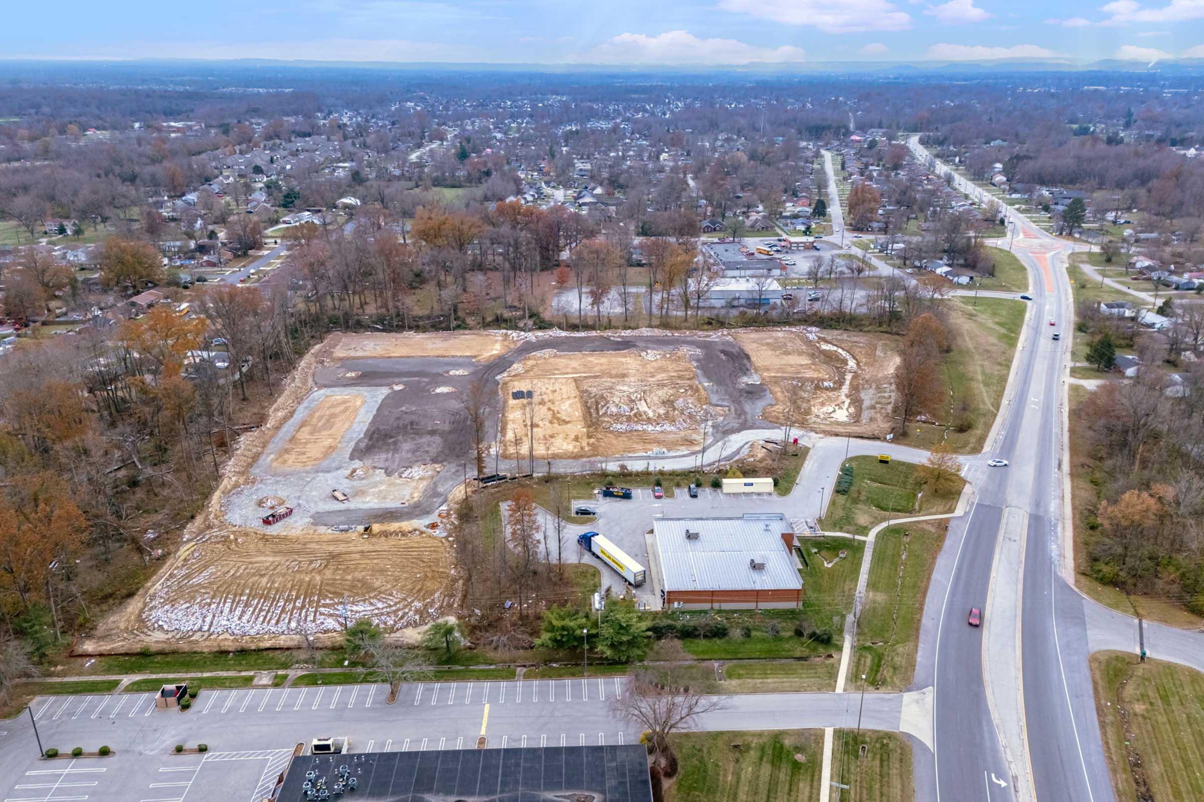 Aerial view of a construction site with cleared land surrounded by trees and a suburban landscape. In the foreground, a parking lot is visible with several parked vehicles. Nearby, a road runs alongside the site, showing light traffic. The scene is characterized by a mix of undeveloped land and residential areas.