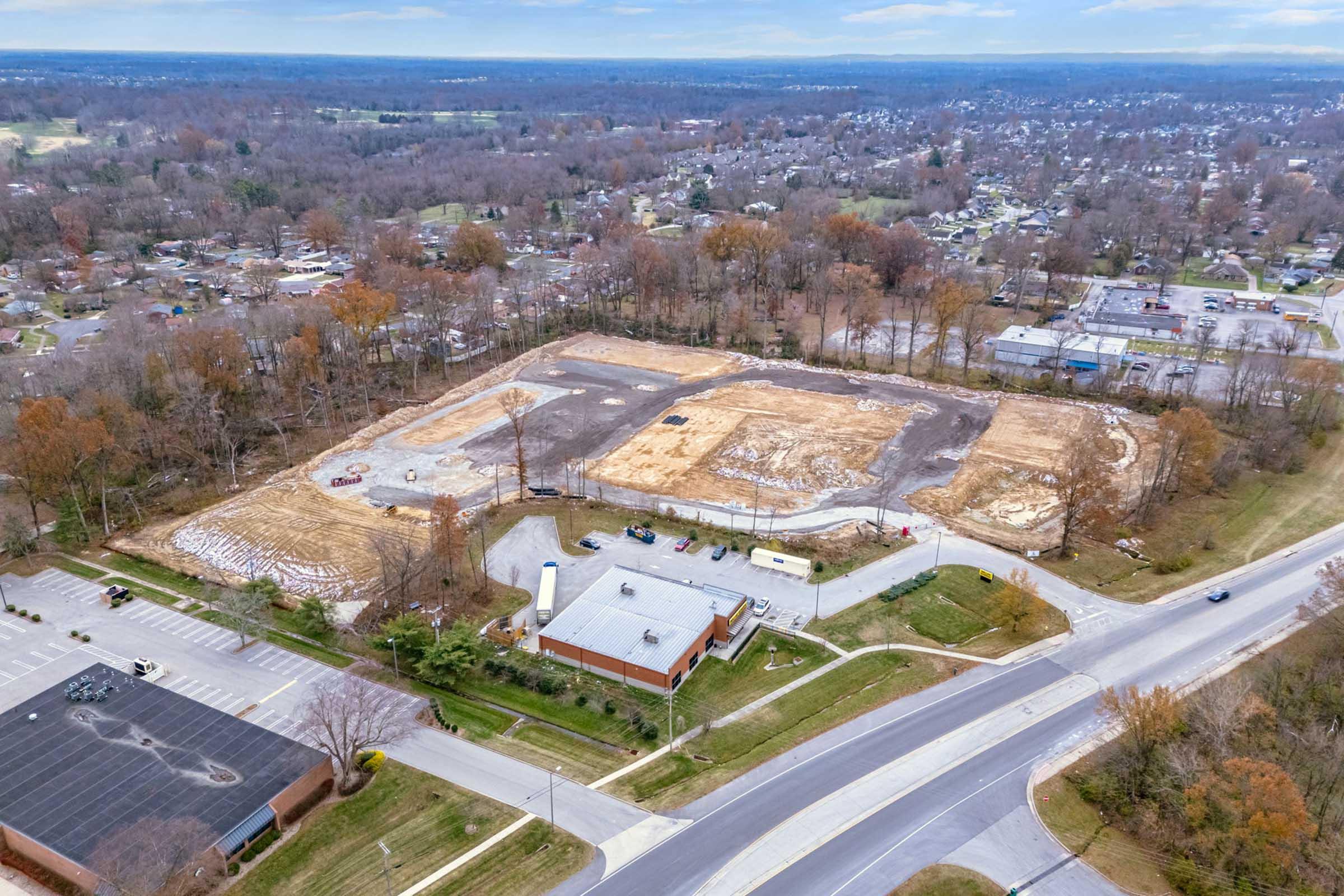 Aerial view of a large, cleared construction site surrounded by trees and residential areas. The site shows bare earth and machinery, with nearby roads and buildings visible, including a parking lot and some structures. The landscape features a mix of rural and suburban elements, with patches of trees remaining.