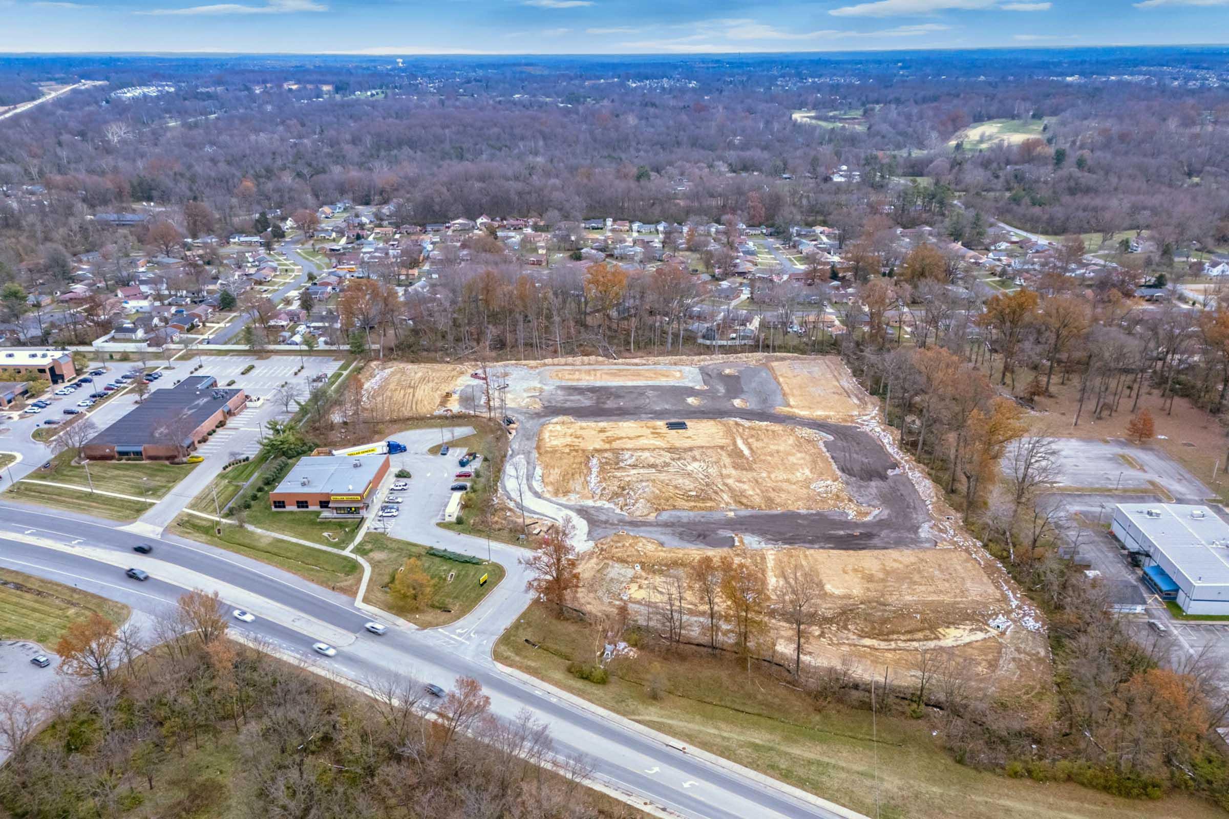 Aerial view of a construction site surrounded by bare trees and residential neighborhoods. The site is cleared, with some areas of gravel and dirt, and nearby roads are visible with vehicles. The landscape shows a mix of developed and natural areas.