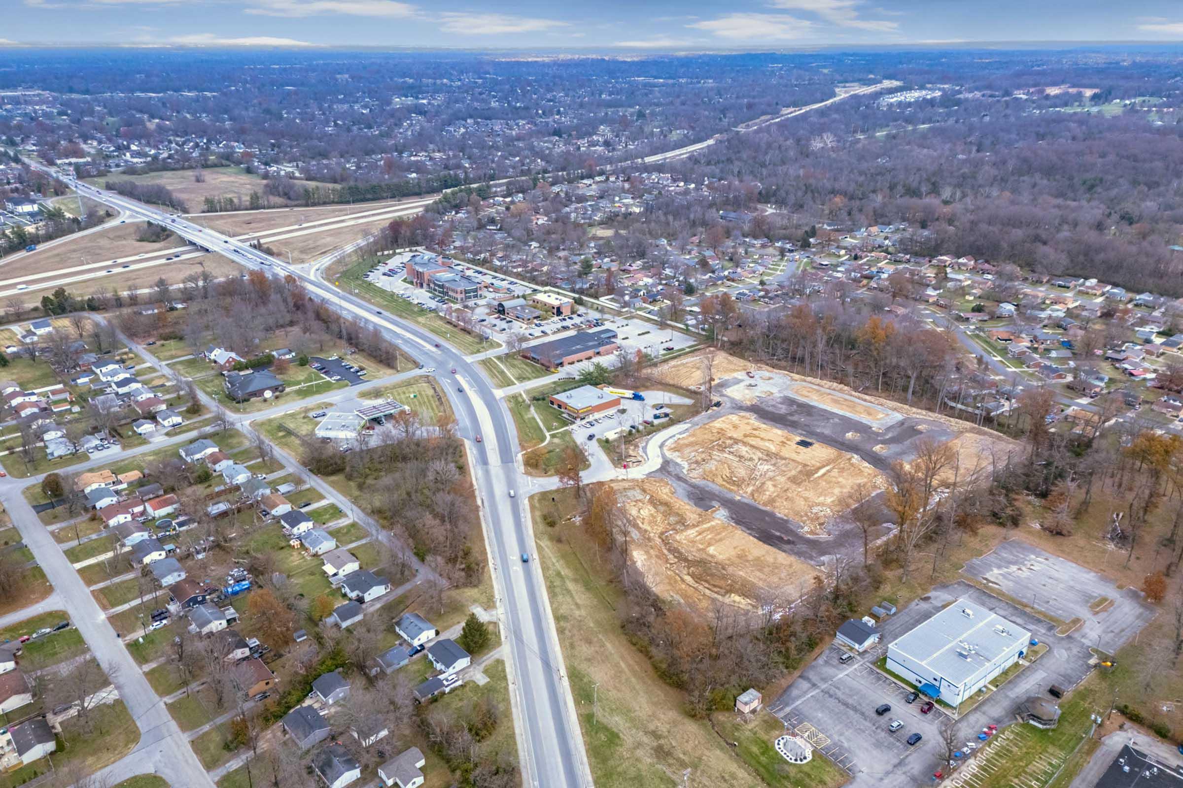 Aerial view of a suburban area showcasing a mix of residential neighborhoods and commercial developments. A major highway runs through the image, with cleared land indicating a construction site. Surrounding the area are trees and houses, reflecting a blend of natural and urban landscapes.