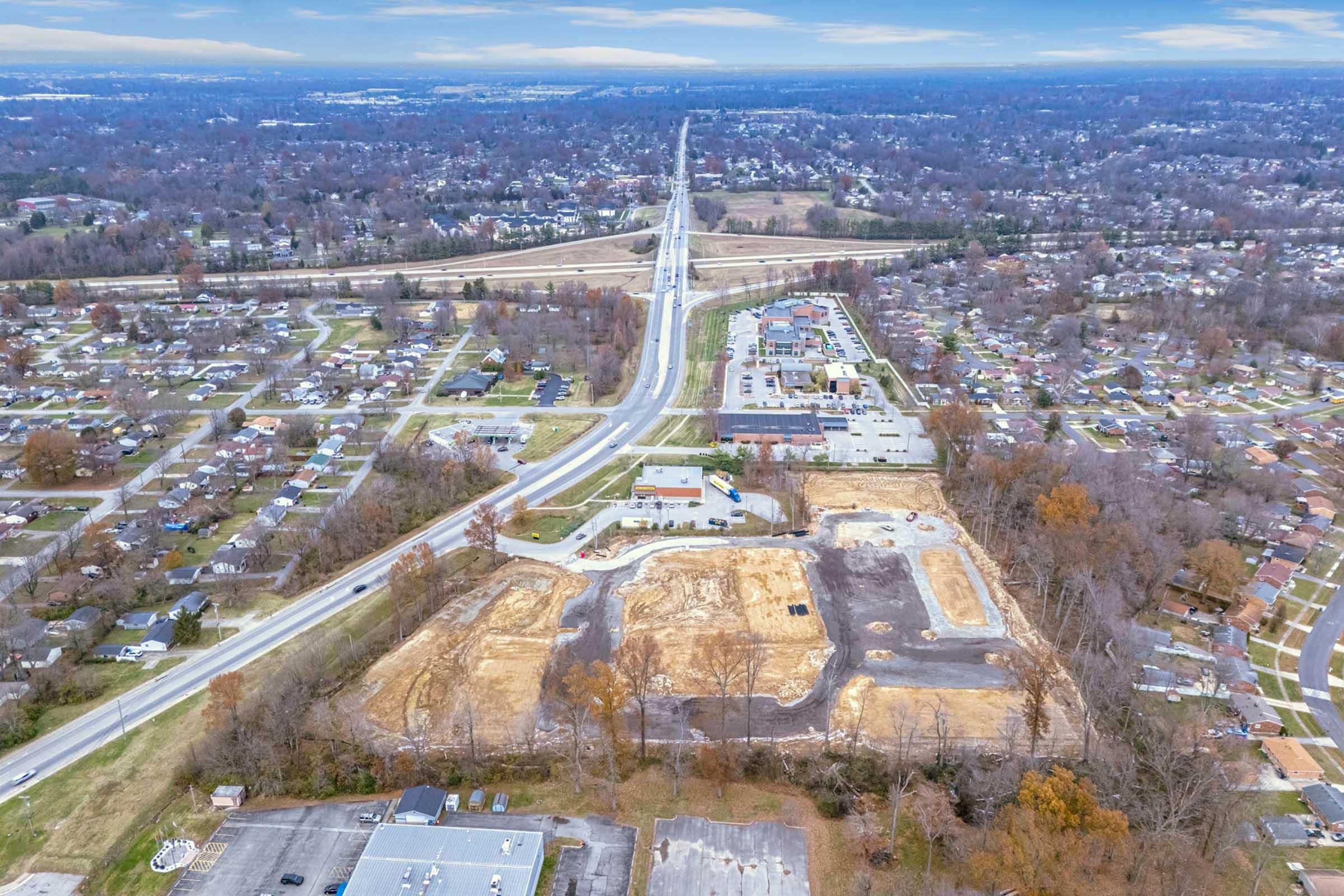 Aerial view of a suburban landscape showing a highway, residential neighborhoods, and cleared land prepared for development. In the foreground, there are construction sites with earth removed, while in the background, there are houses and commercial buildings along the road. The sky is overcast.