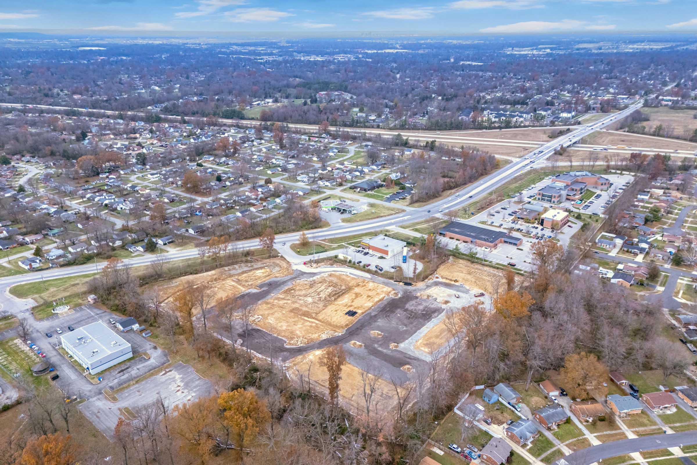 Aerial view of a suburban area showing construction sites and cleared land. Surrounding residential neighborhoods, roads, and patches of trees are visible. In the distance, a highway runs through the landscape, with various buildings and infrastructure in the vicinity. The scene is set against a cloudy sky.