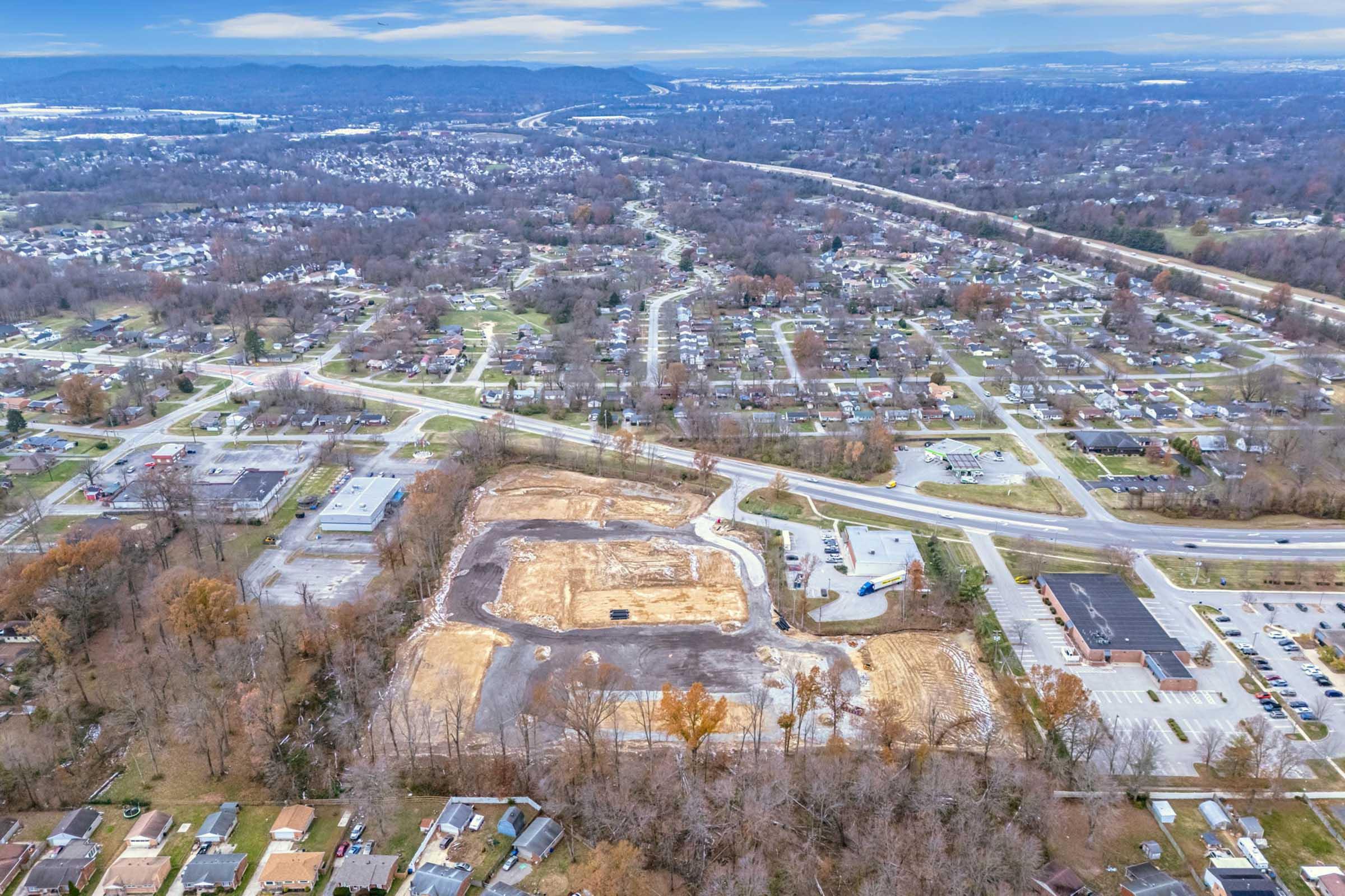 Aerial view of a suburban area showing a mix of cleared land and developed neighborhoods. The foreground features a large, cleared plot, while the background displays houses and a grid of roads. Trees with autumn foliage are visible, and a highway runs through the landscape.