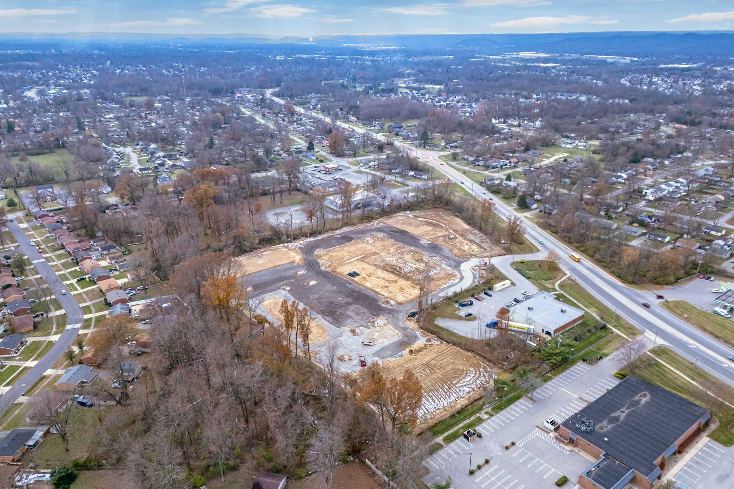 Aerial view of a construction site surrounded by residential areas and trees, showing cleared land with earth-moving equipment. Nearby roads are visible, along with a parking lot and several houses in the background, indicating urban development. The landscape includes patches of autumn foliage.