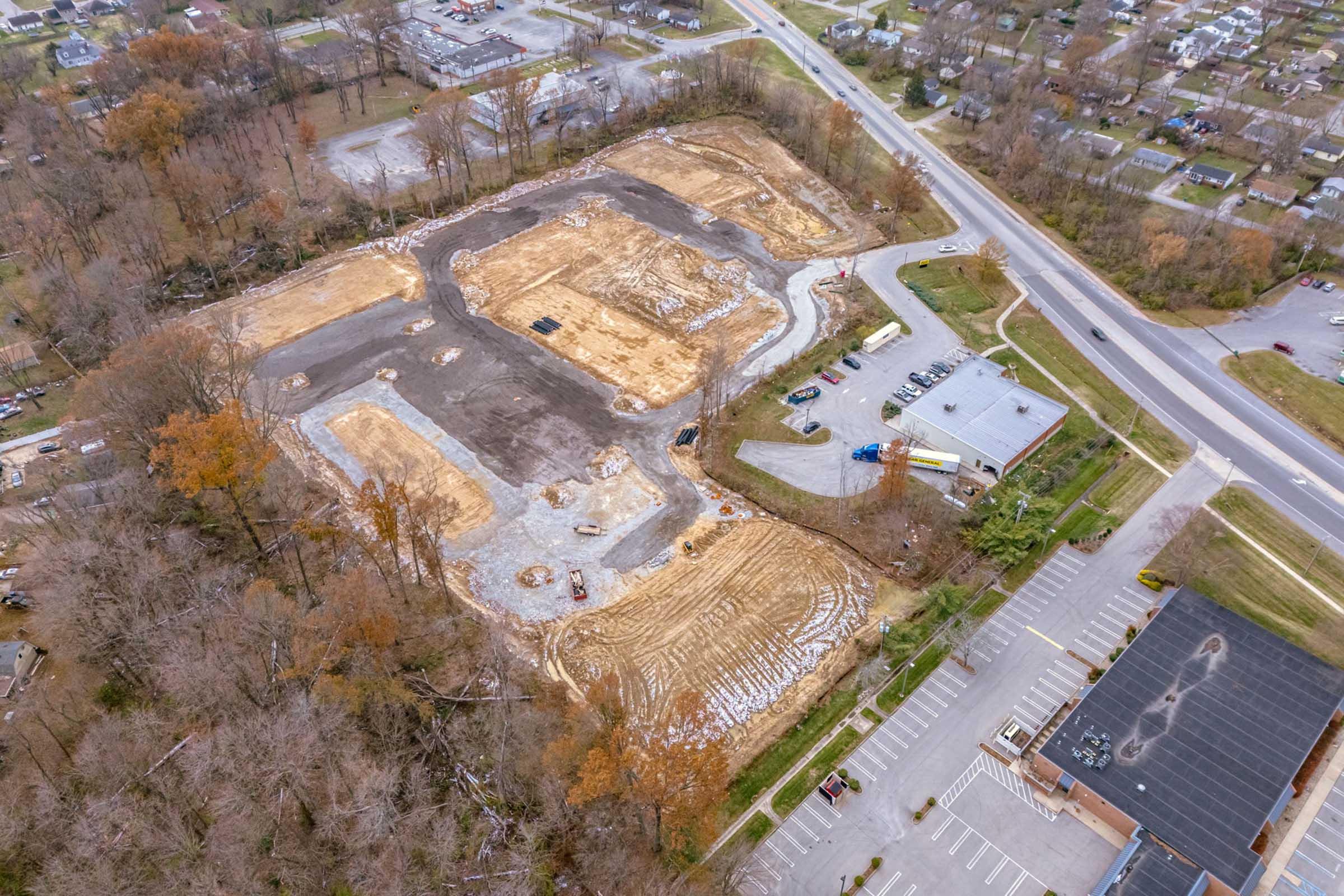Aerial view of a large construction site with cleared land showing dirt and gravel, surrounded by trees with some autumn foliage. Nearby, a paved parking area is visible, as well as a road leading to residential buildings in the background. The area appears to be in the early stages of development.
