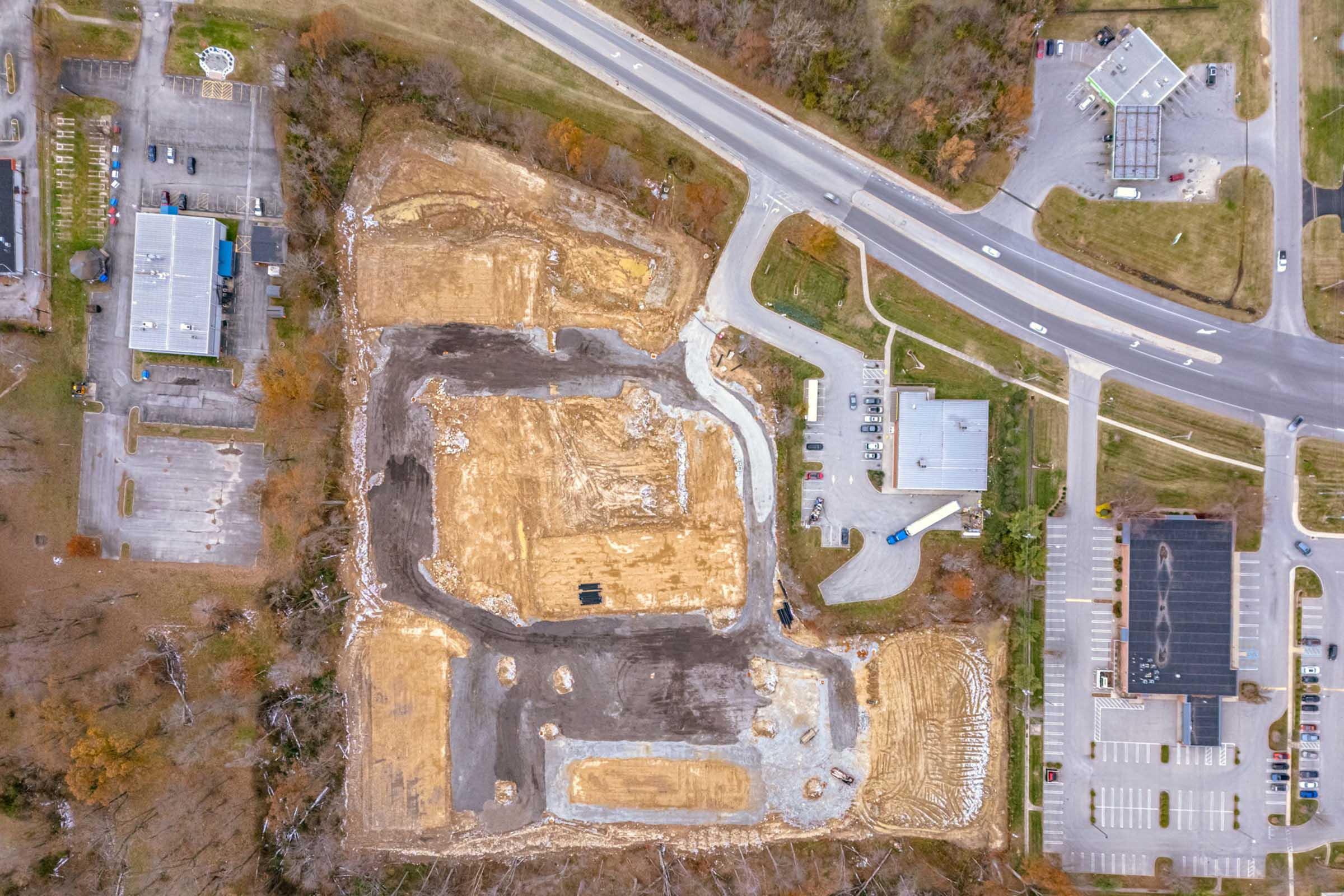 Aerial view of a construction site with cleared land and excavation areas, surrounded by trees and roads. Nearby are parking lots and commercial buildings. The site features a mix of dirt, gravel, and paved areas, indicating ongoing development.