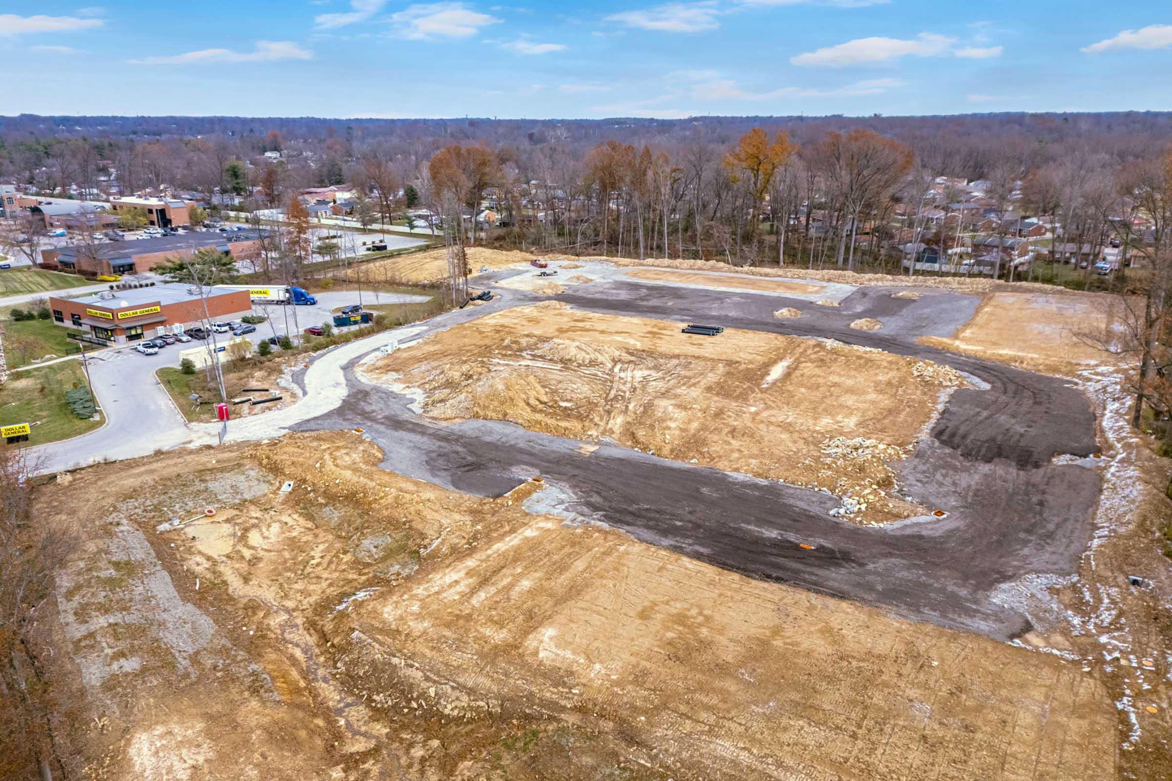 Aerial view of a construction site with cleared land and minimal vegetation. Gravel and dirt fill the area, with scattered rocks and machinery visible. In the background, a few suburban buildings and trees can be seen, along with a fast-food restaurant on one side of the image. The sky is partly cloudy.