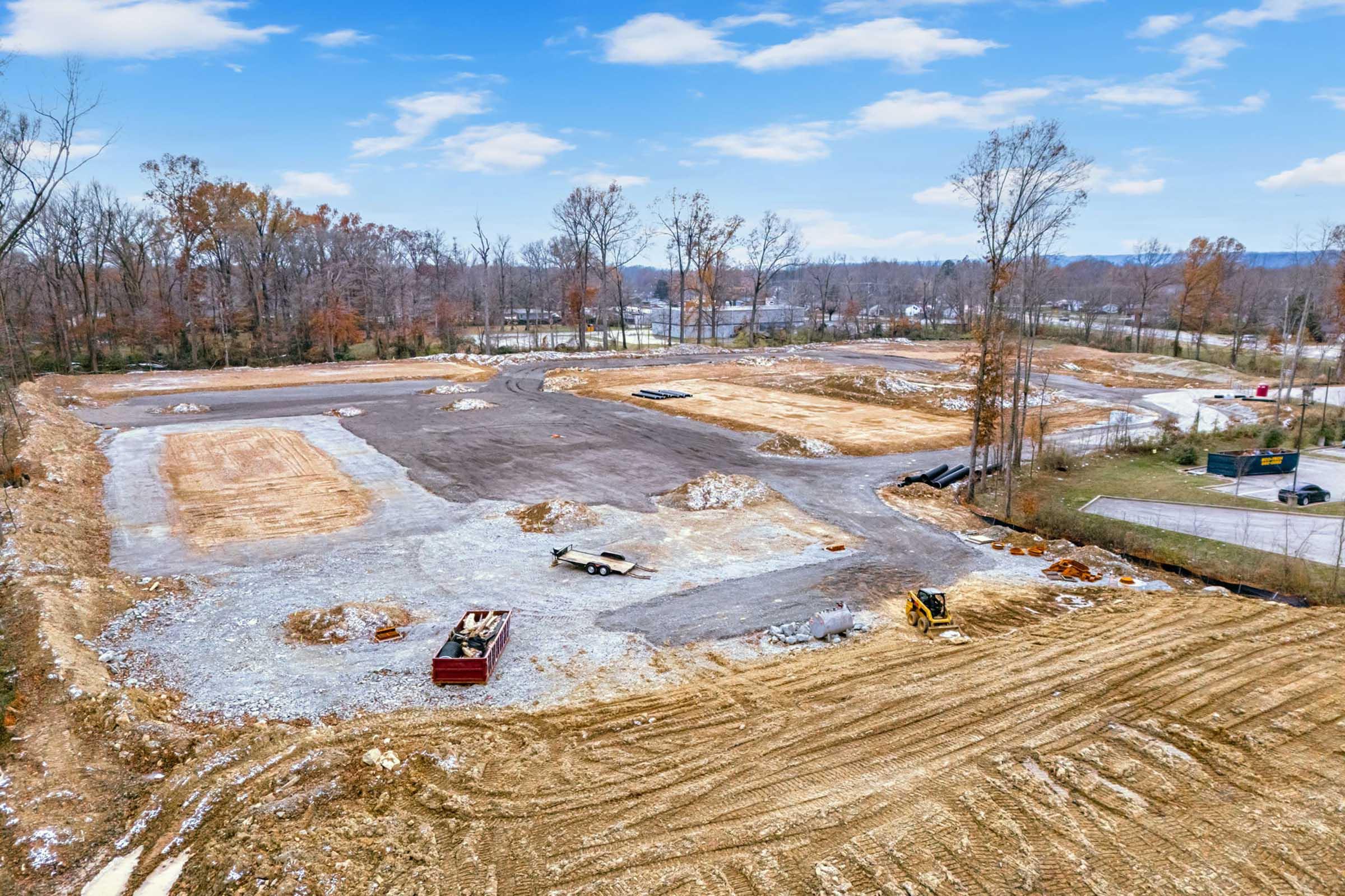 Aerial view of a construction site featuring cleared land with exposed soil and gravel. Various machinery and equipment are visible, alongside piles of dirt and construction materials. Trees in the background appear bare, indicating a winter season. The overall scene depicts early stages of development.