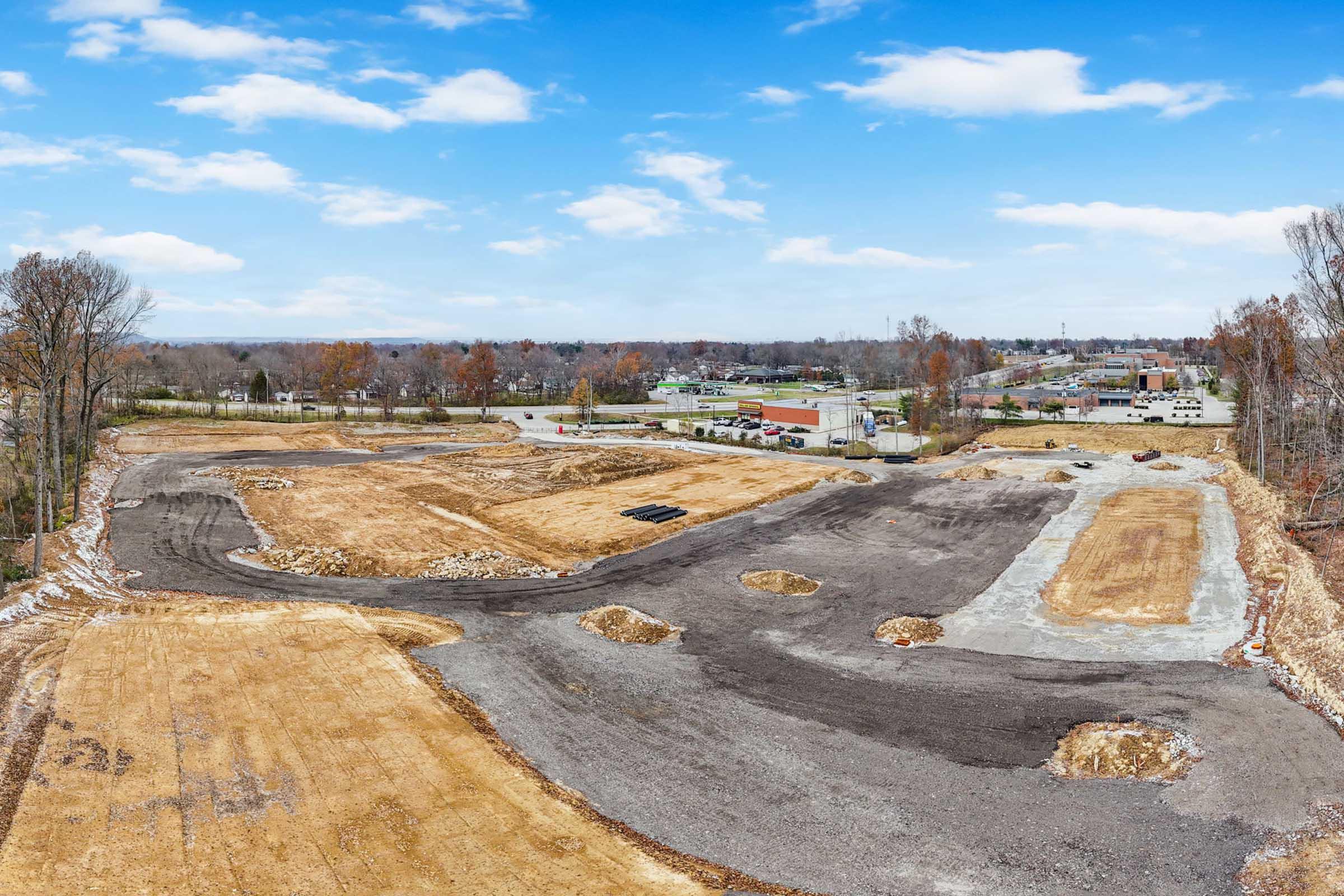 A wide view of a cleared construction site with a mix of bare earth, gravel, and patches of vegetation. The landscape is dotted with piles of dirt and rocks, with a suburban area visible in the background, including buildings and trees under a clear blue sky with scattered clouds.