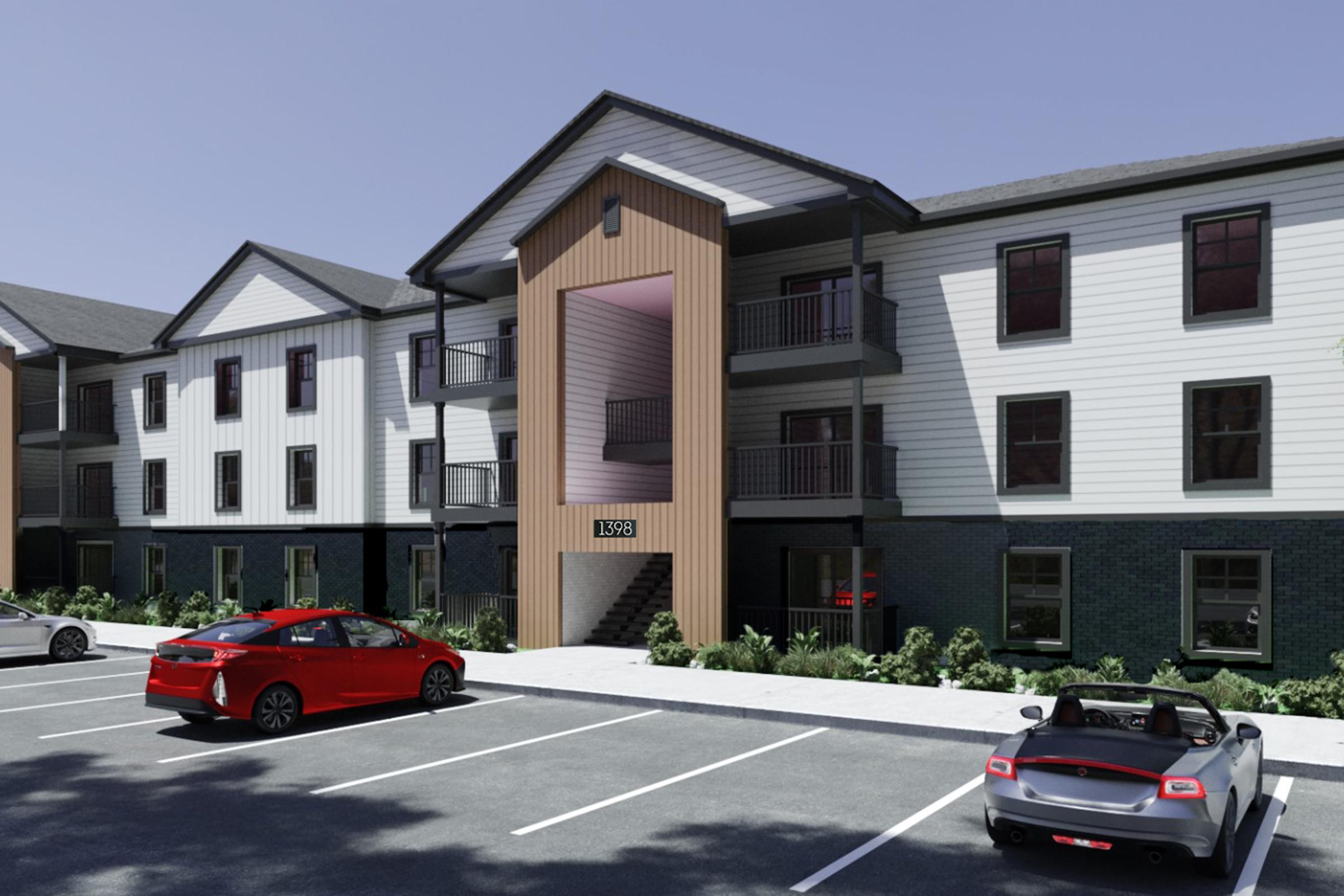 Exterior view of a modern multi-unit residential building with a mix of white and dark brick siding. Features include a central entrance with wooden accents, balconies on the upper floors, and a parking lot with red and silver cars parked in front. Lush landscaping surrounds the building.
