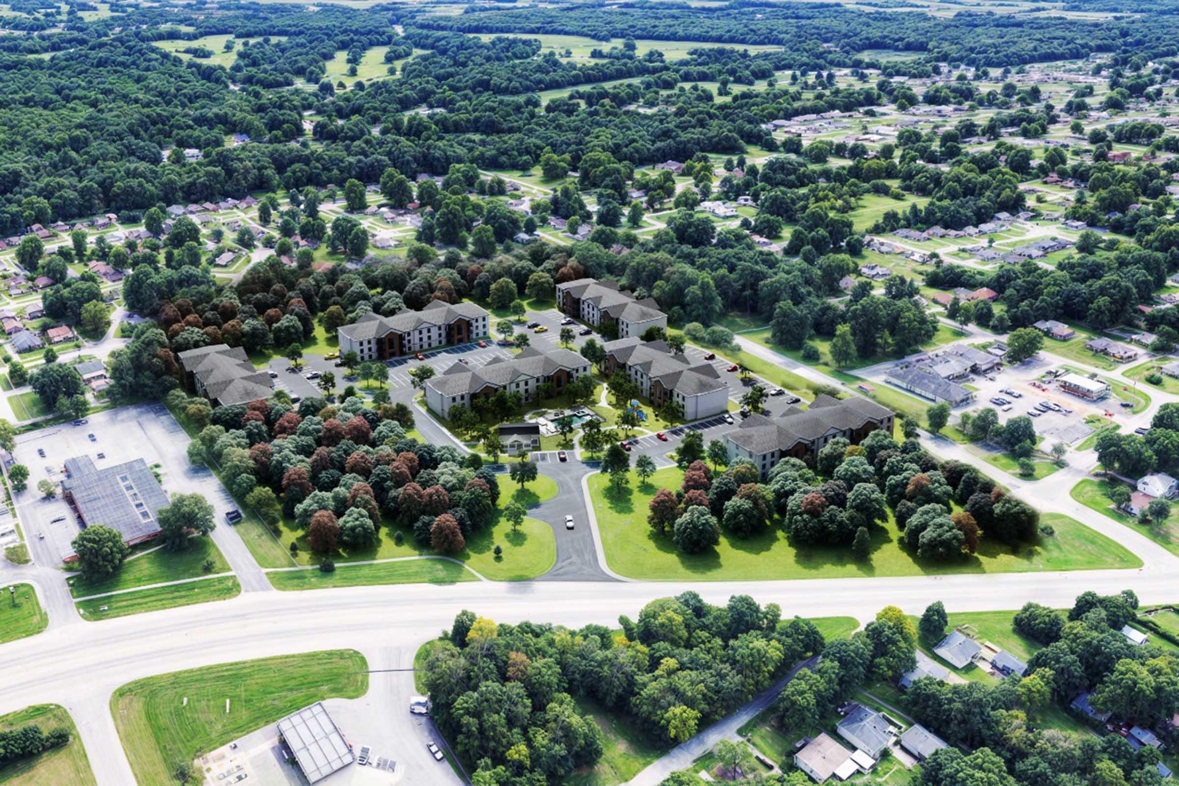 Aerial view of a large residential complex surrounded by greenery, featuring multiple buildings with varied architectural styles. The area is dotted with trees in autumn colors and is adjacent to a road, with additional buildings and parking spaces visible nearby. Lush fields and hills extend in the background.