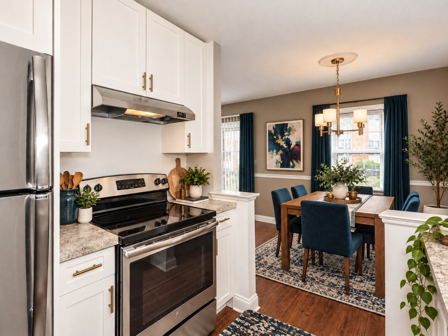 A modern kitchen with stainless steel appliances, white cabinets, and a granite countertop. In the background, a dining area features a wooden table surrounded by blue upholstered chairs, a chandelier overhead, and large windows with blue curtains. Decorative plants and artwork add warmth to the space.