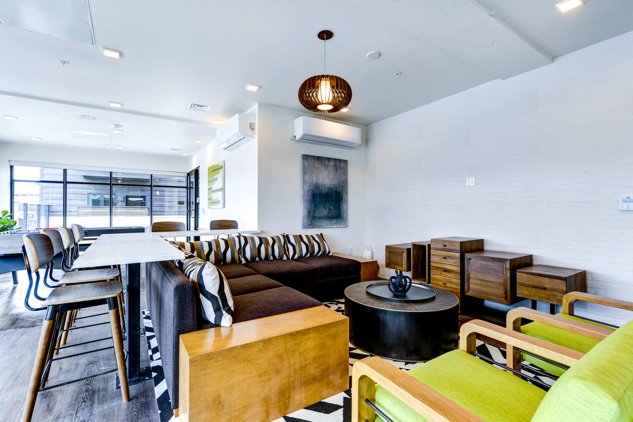 Stylish modern lounge area featuring a sectional sofa with striped cushions, a round coffee table, and sleek wooden furniture. The decor includes a pendant light, air conditioning units, and large windows providing natural light. The space is designed with a contemporary aesthetic and a black-and-white patterned rug.