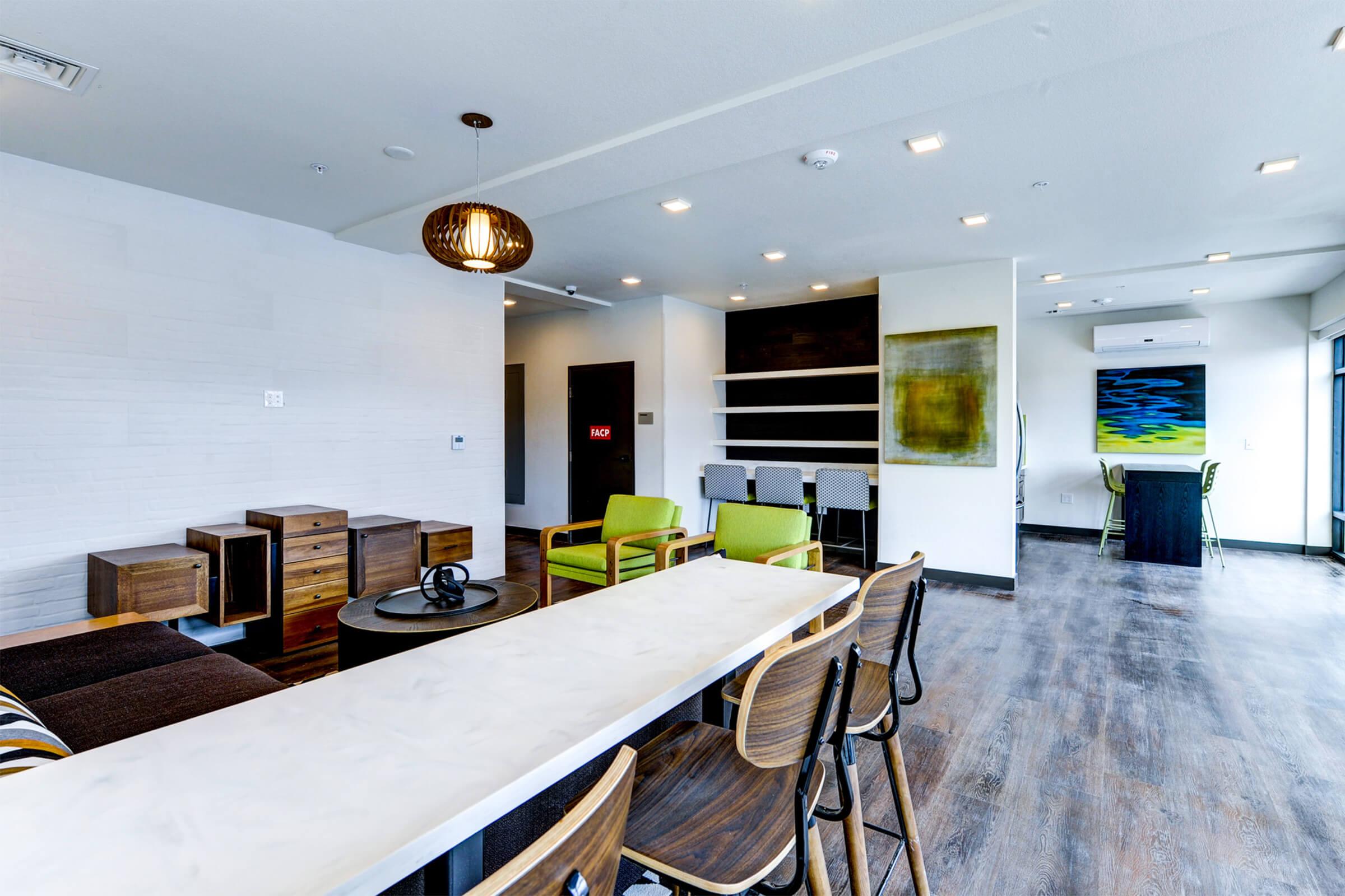 A modern communal lounge featuring a long white table, wooden chairs, storage units, and a vibrant green and blue artwork on the wall. The space is well-lit with pendant lighting, showcasing a contemporary design with a mix of textures and materials.