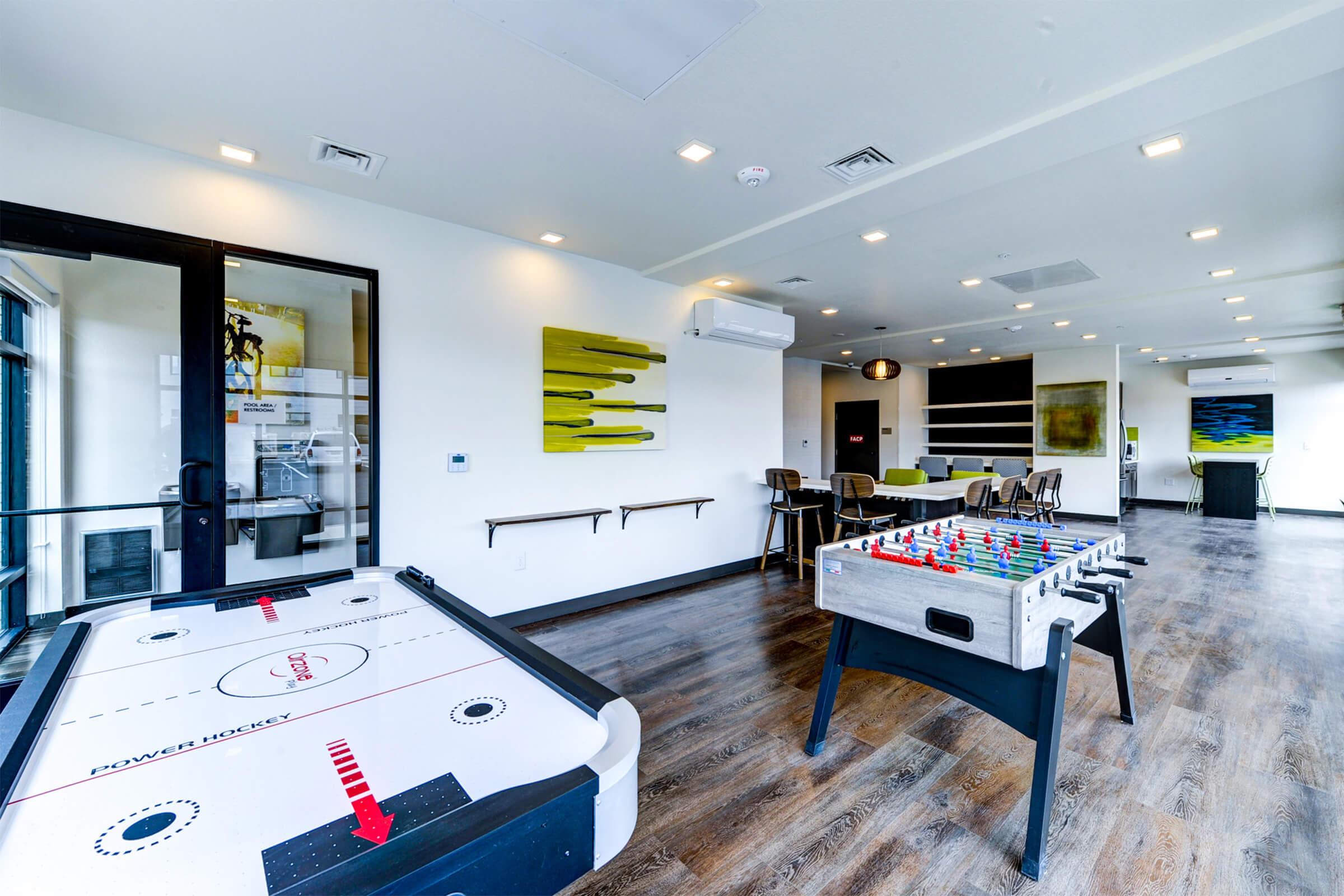 A modern game room featuring an air hockey table and a foosball table. The space has light wood flooring, bright overhead lighting, and contemporary furniture. There are large windows along one wall, and the decor includes abstract artwork and a vibrant color palette.