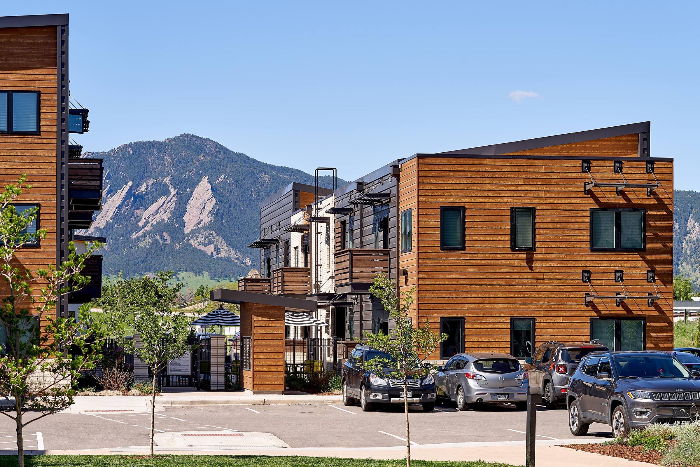 Modern multi-story buildings with wooden exteriors, situated in a scenic area. In the foreground, several parked cars are visible, along with landscaped greenery. In the background, majestic mountains rise under a clear blue sky, creating a picturesque atmosphere.